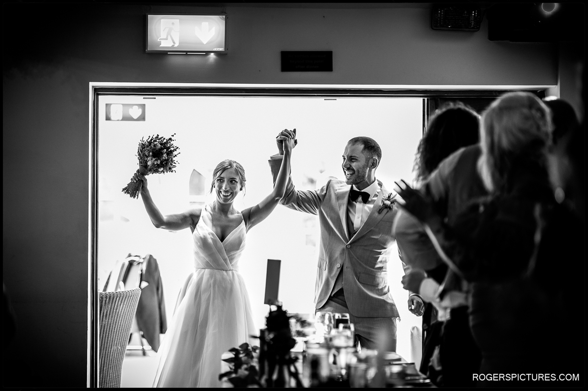 Bride and groom entering the reception holding hands in the air as guests applaud.