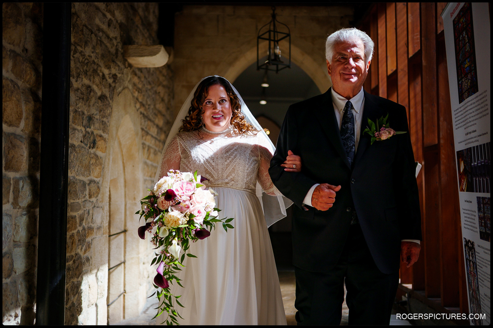 Bride walks arm-in-arm with her father through the historic stone hallway at St Etheldreda’s Church, holding a cascading floral bouquet in soft pinks and burgundy tones.