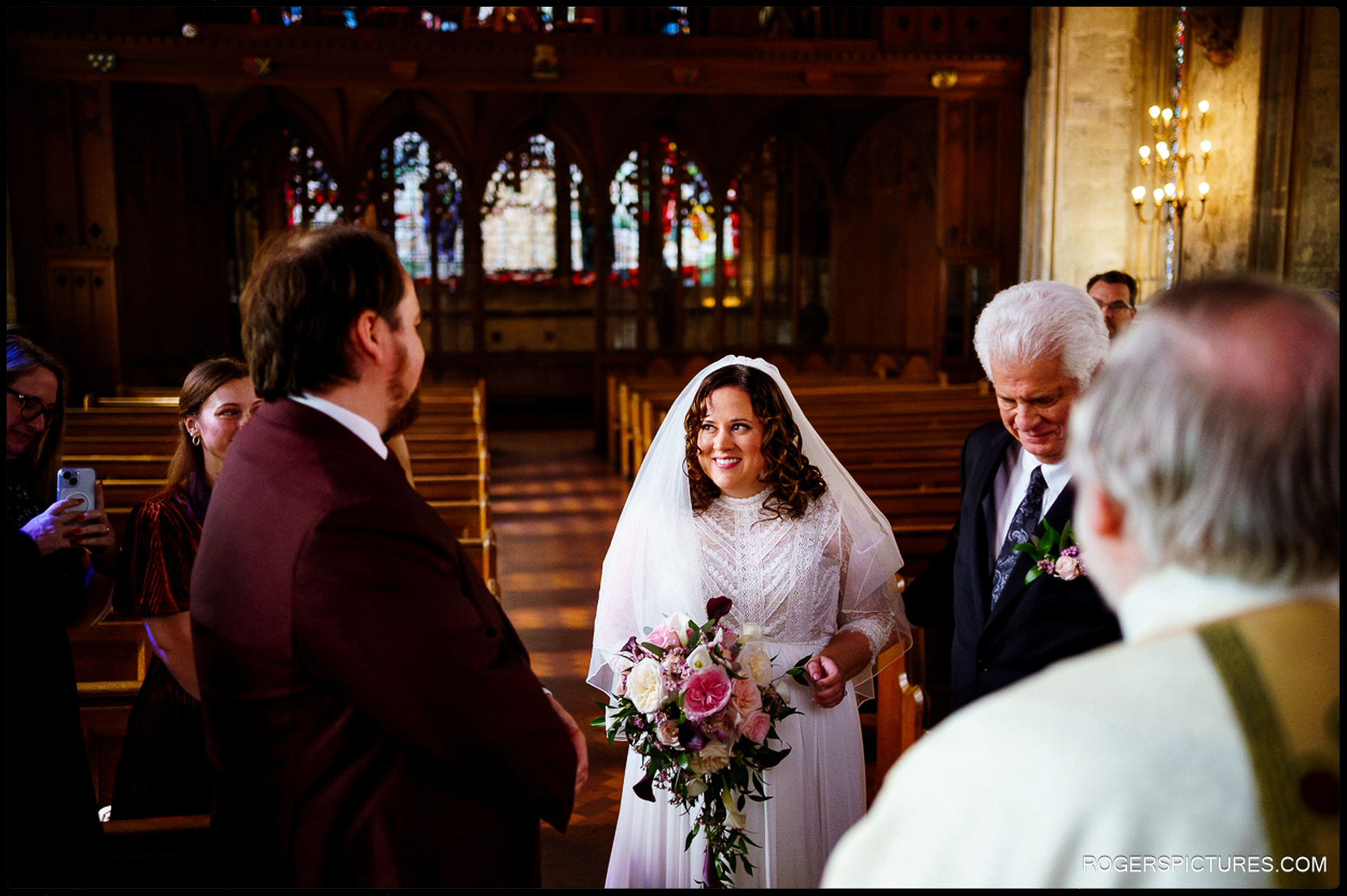 Bride smiles at groom as her father walks her down the aisle inside St Etheldreda’s Church, natural candid moment during the ceremony.