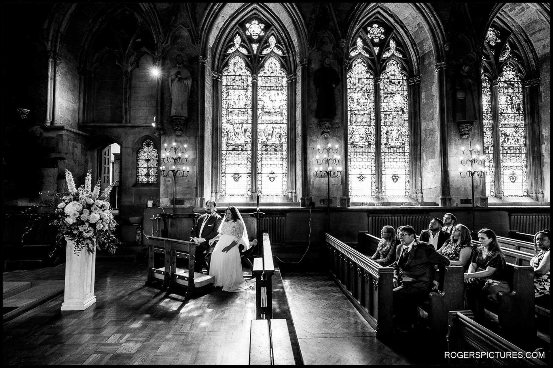 Bride and groom sit together at the front of St Etheldreda’s Church during the Nuptial Mass, dramatic gothic arches and tall stained-glass windows behind them.