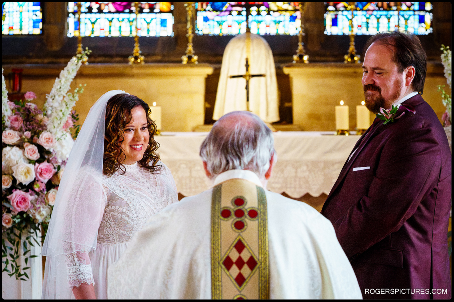 Bride smiles warmly at the priest while standing beside the groom at the altar of St Etheldreda’s Church, surrounded by flower arrangements and candlelight.