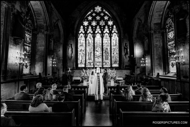 Destination London St Etheldreda’s Church Wedding 015 Wide, black-and-white view of wedding ceremony inside St Etheldreda’s Church, showing the couple at the altar beneath towering gothic windows while guests sit in pews.