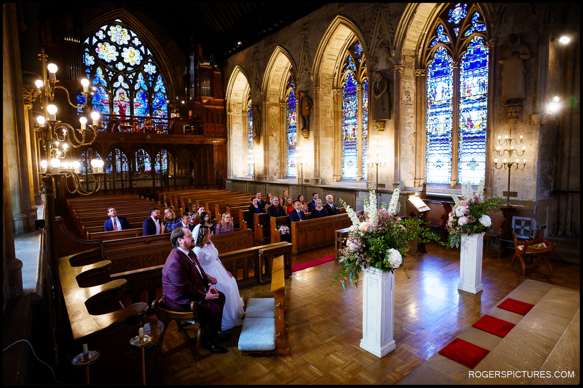 Guests sitting in pews watching the couple during their wedding ceremony at St Etheldreda’s Church, colourful stained-glass windows and chandeliers in the background.
