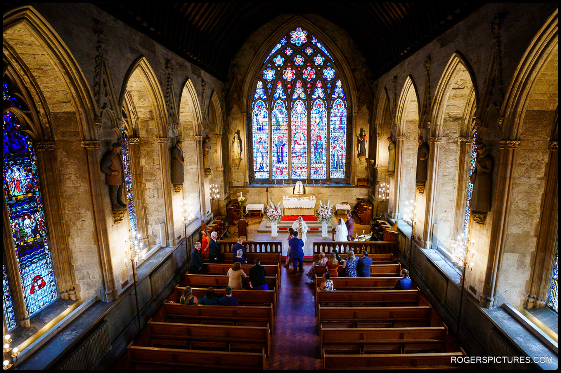 Wide view of wedding ceremony inside St Etheldreda’s Church from the balcony, showing the couple at the altar beneath large stained-glass windows and gothic arches.