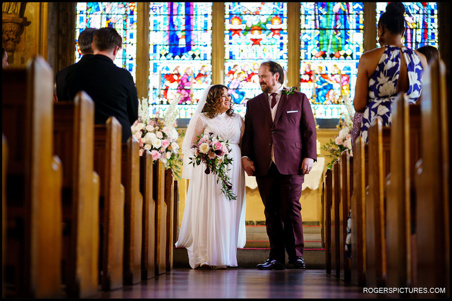Bride and groom walk hand-in-hand down the aisle at St Etheldreda’s Church, smiling at each other with colourful stained-glass windows behind them.