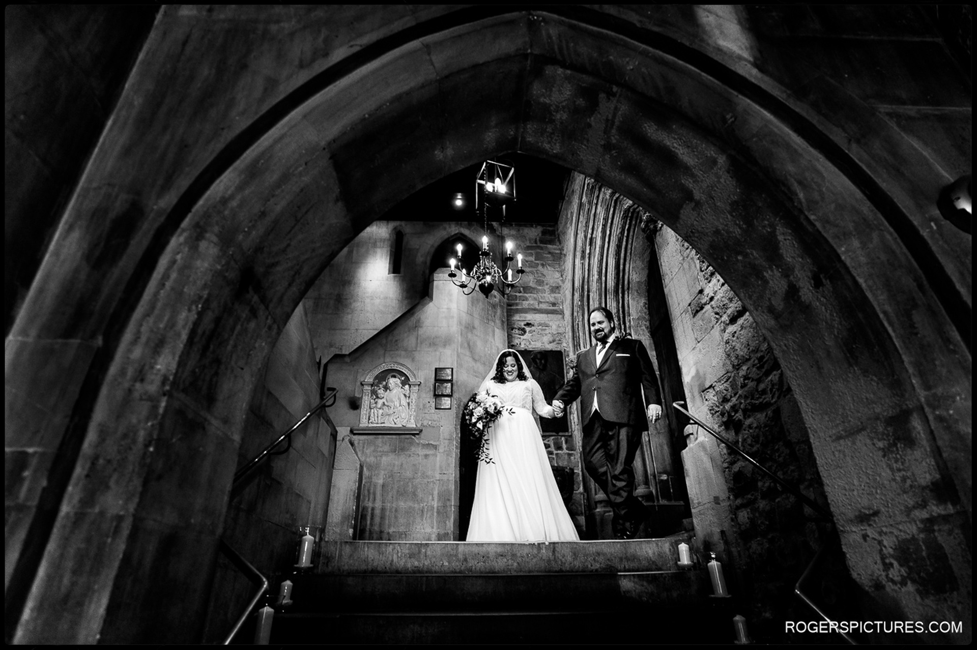 Bride and groom walk down a stone staircase beneath a dramatic gothic arch inside St Etheldreda’s Church, holding hands after the ceremony.
