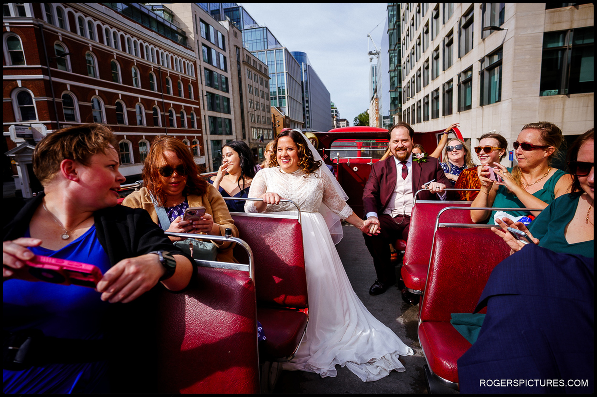 Bride and groom sit together on an open-top red London bus with guests, laughing and taking in the city during their wedding day tour.