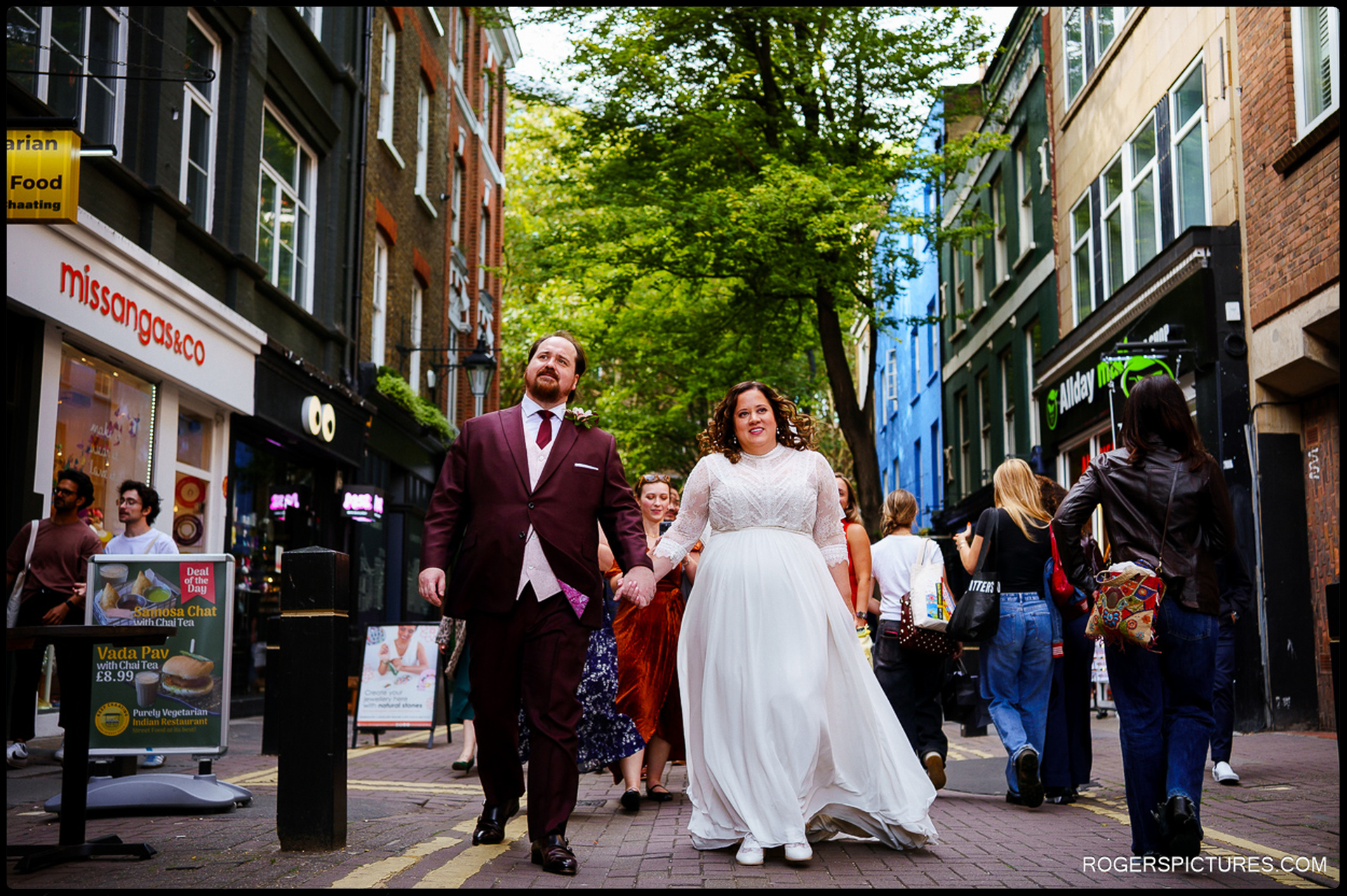 Bride and groom walk through vibrant London streets hand-in-hand with guests behind them, blending into city life after the ceremony.