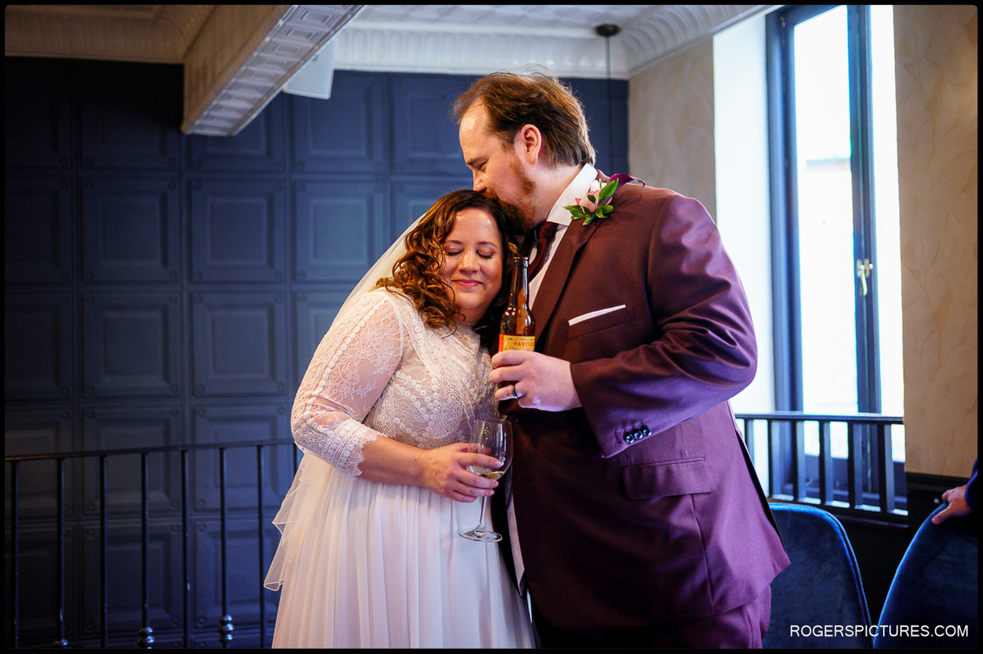 Bride leans into groom for a quiet embrace during drinks reception, holding a wine glass as the groom gently kisses her forehead.