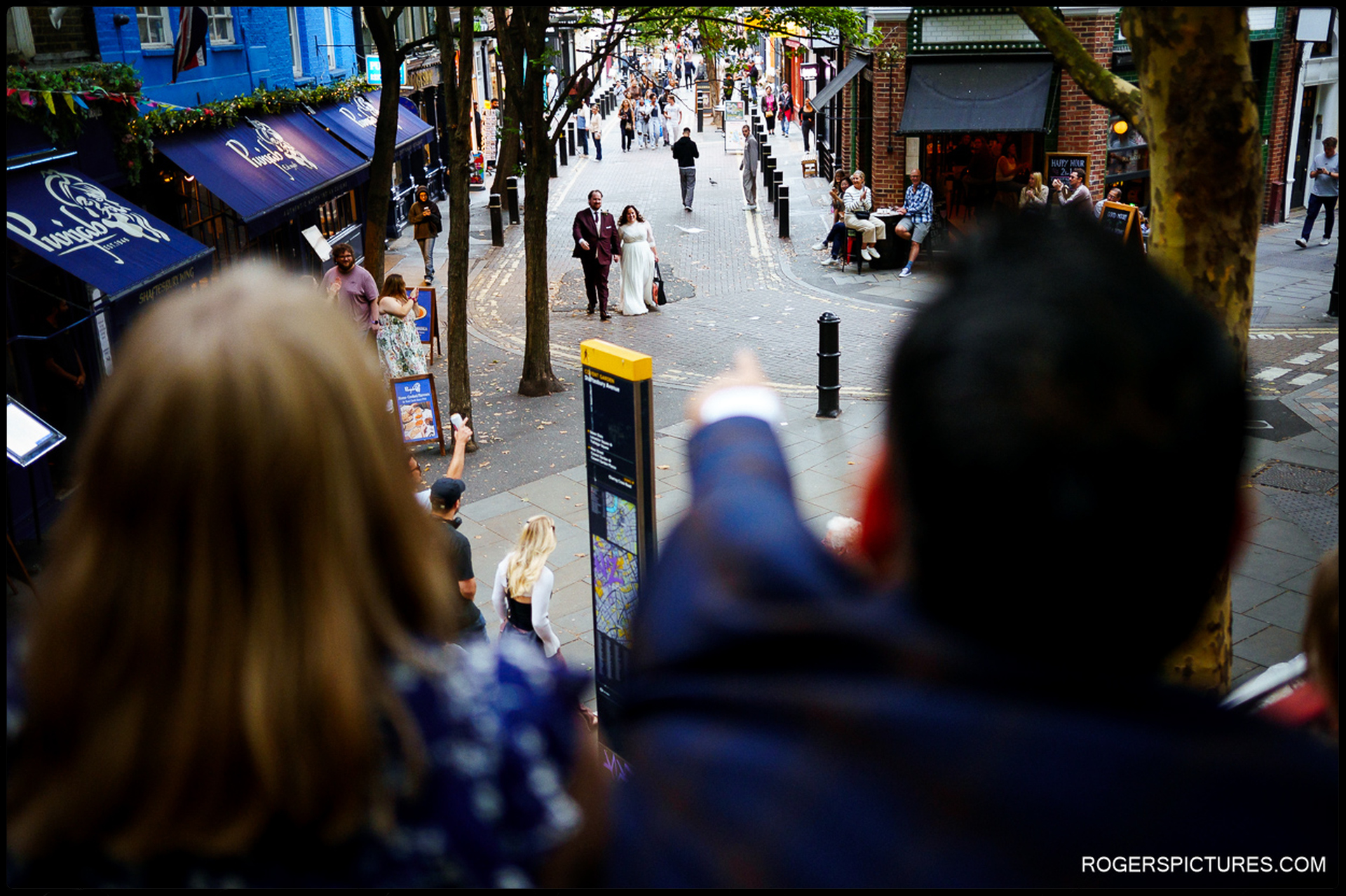 Guests on an open-top bus excitedly point as the bride and groom walk along a busy London street below, surrounded by cafés and passers-by.