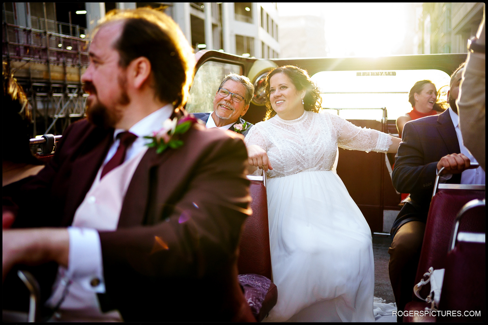Bride smiles in golden evening light while riding an open-top bus through London with the groom and guests, relaxed and enjoying the city views.