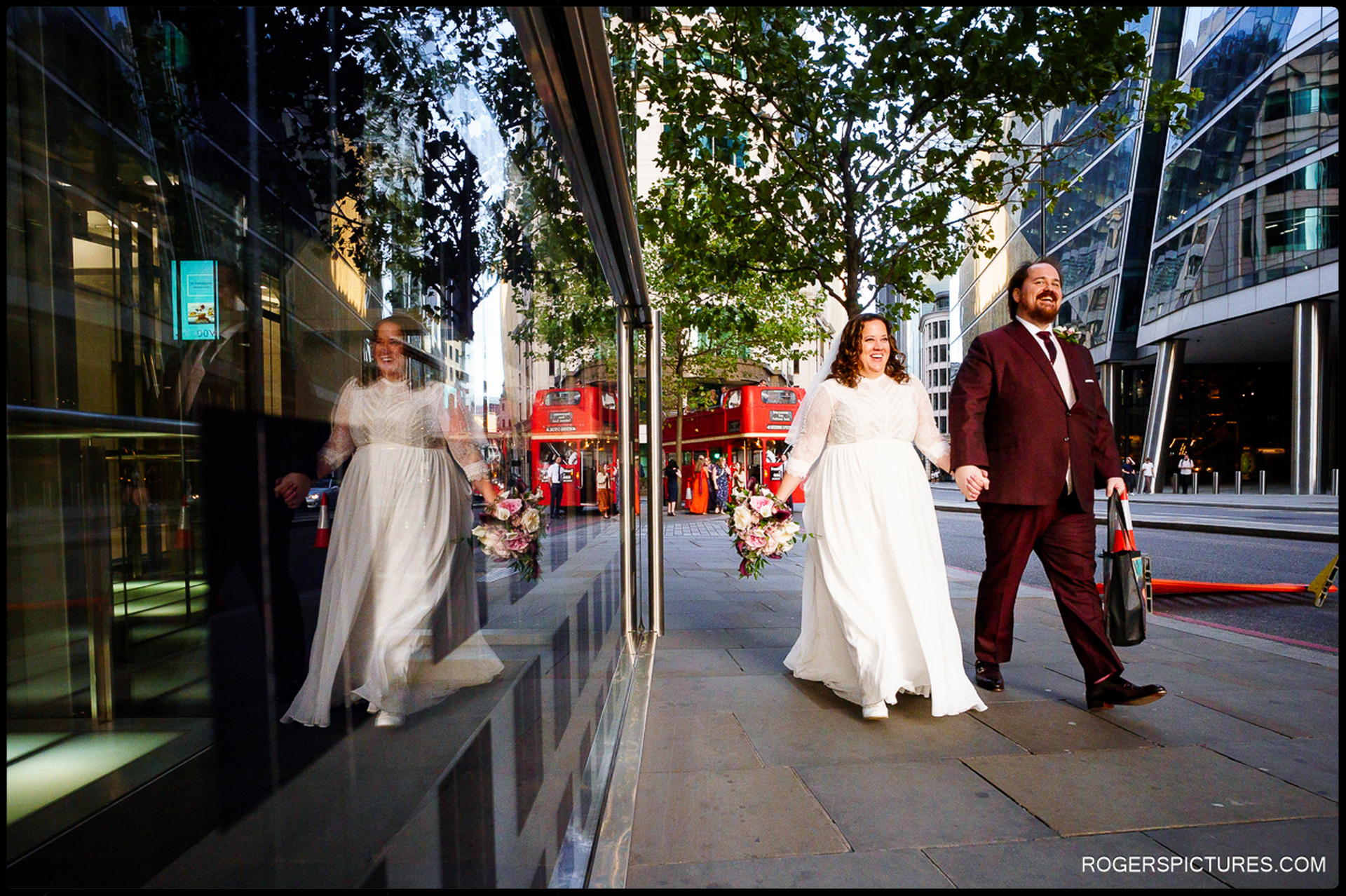 Bride and groom walk hand-in-hand through central London after their wedding, reflected in a glass building with classic red double-deckers in the background.
