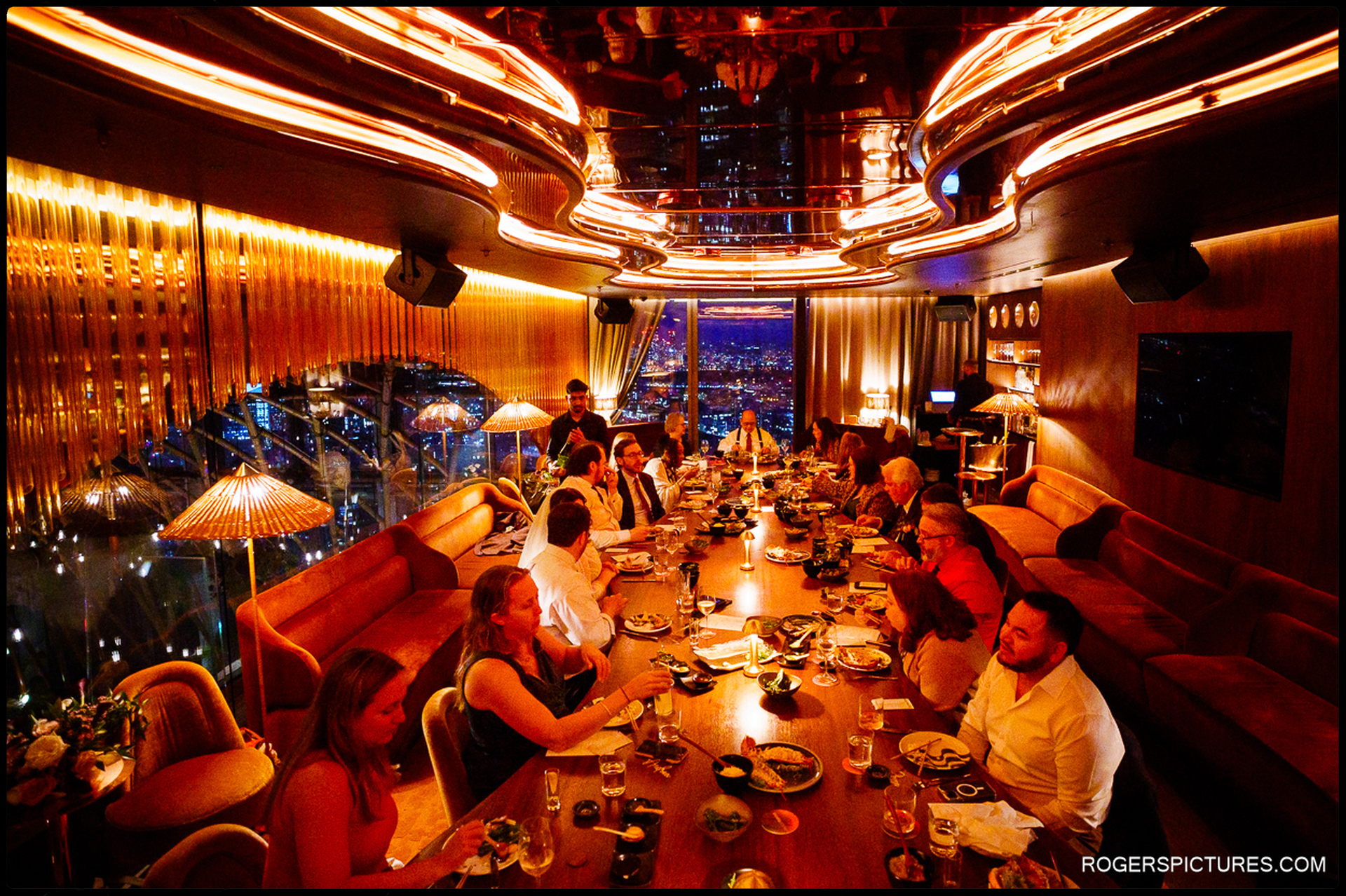 Wedding guests enjoying dinner in the private dining room at SushiSamba, glowing orange lighting and city skyline visible through floor-to-ceiling windows.