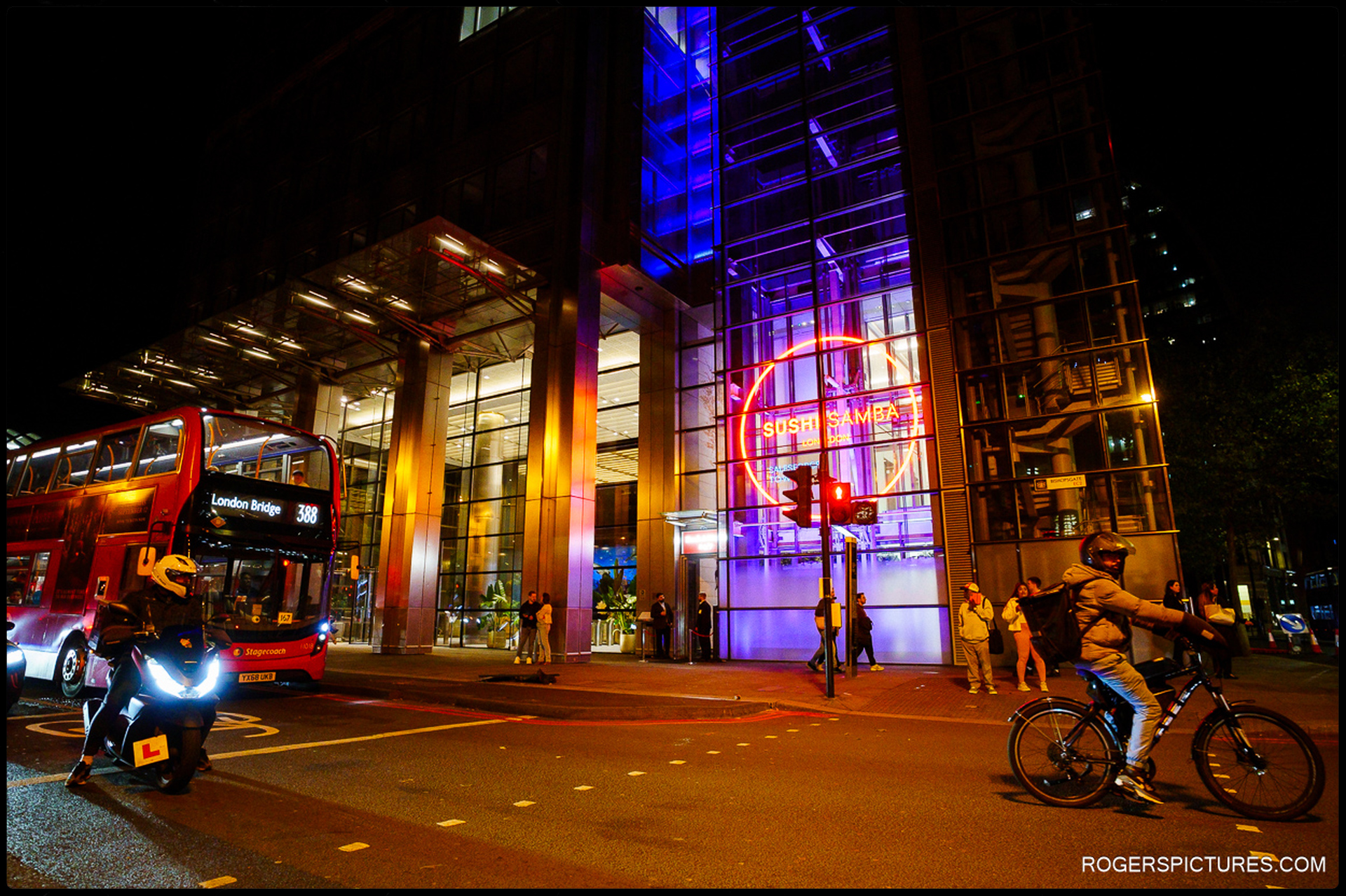 Exterior night view of SushiSamba London with neon signage glowing, red London bus and motorbike passing by on the street.