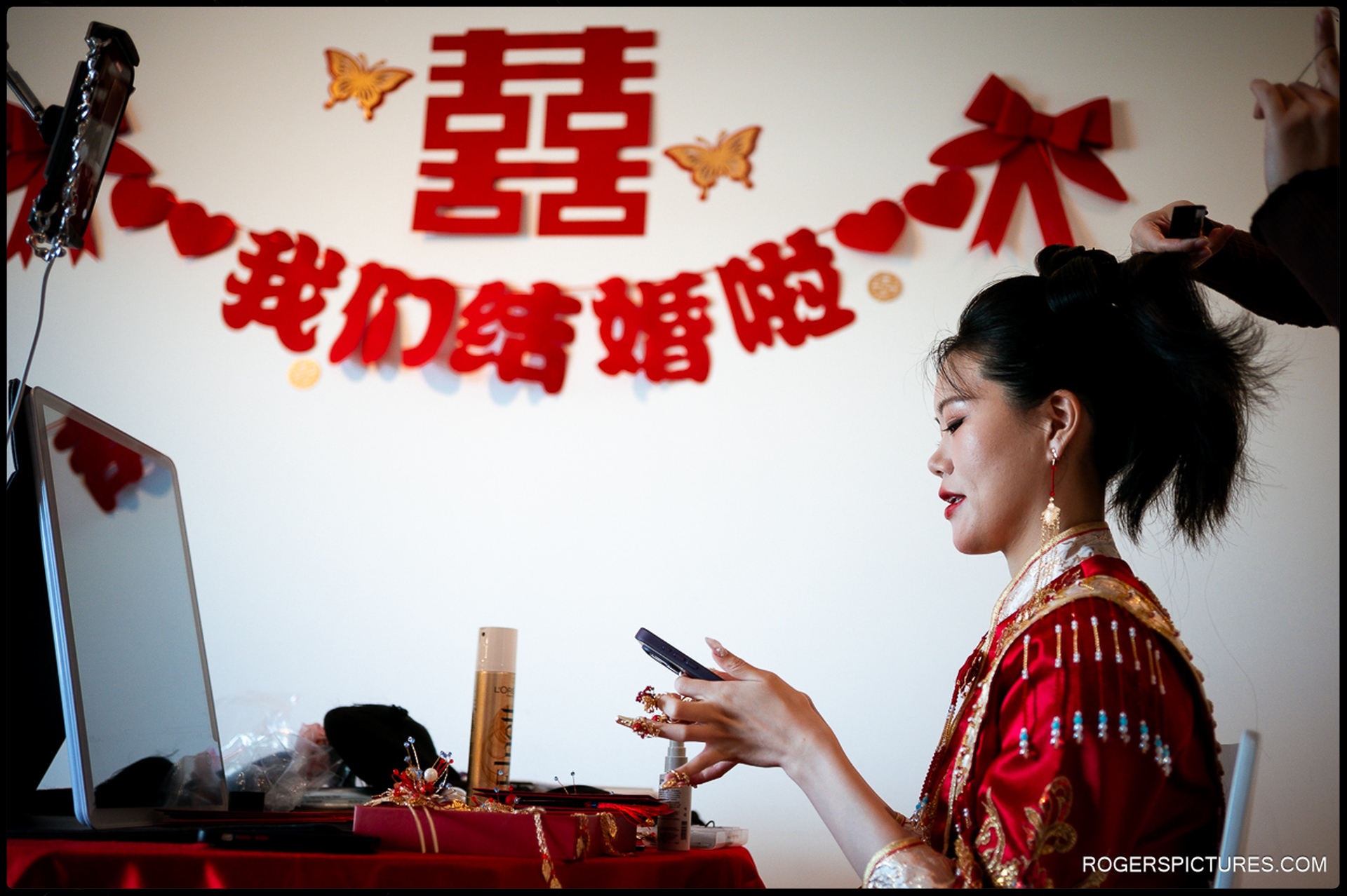Bride having her hair styled while sitting at a dressing table decorated with red Chinese wedding symbols