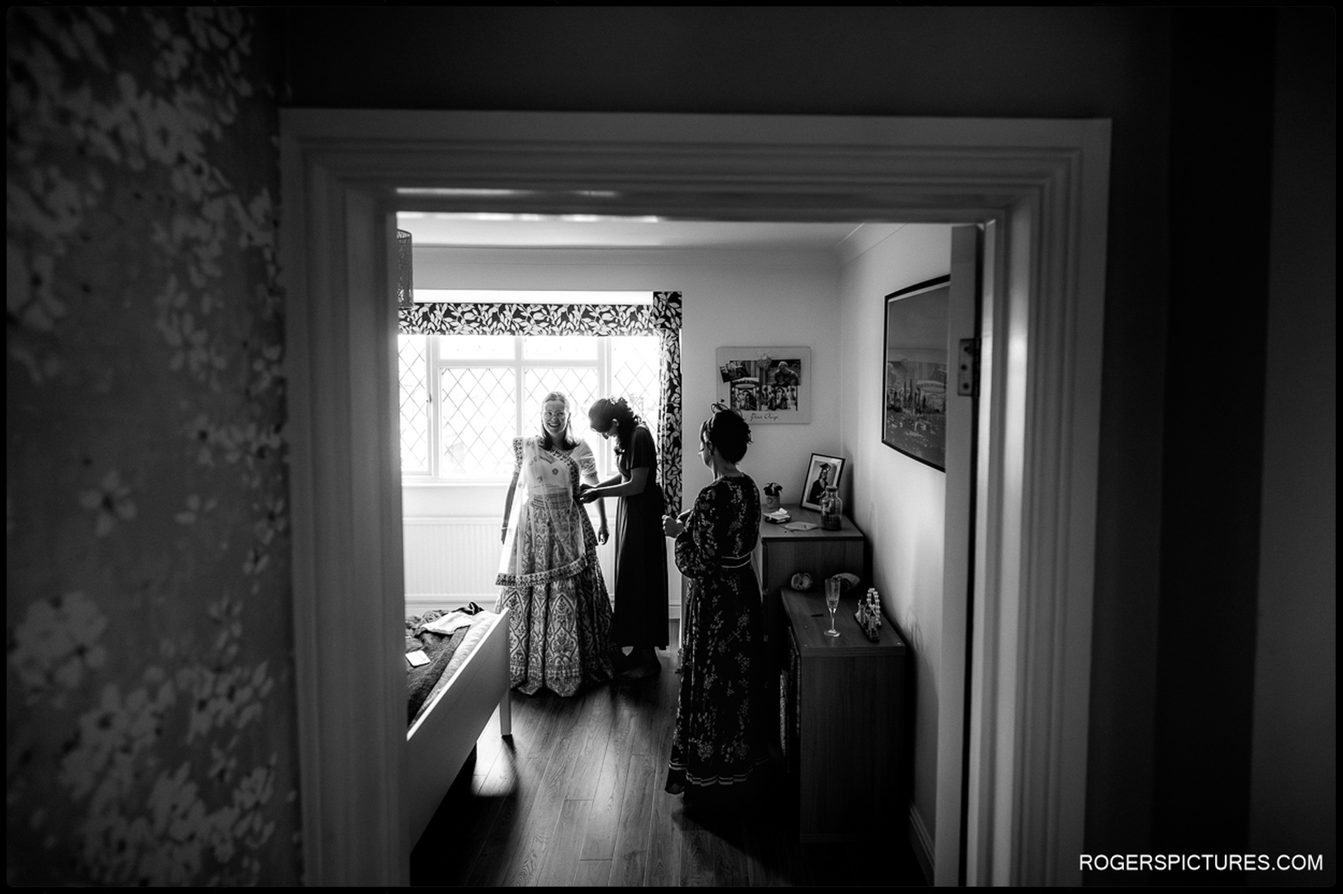 Bride stands in a bright bedroom while two bridesmaids help adjust her embroidered outfit.