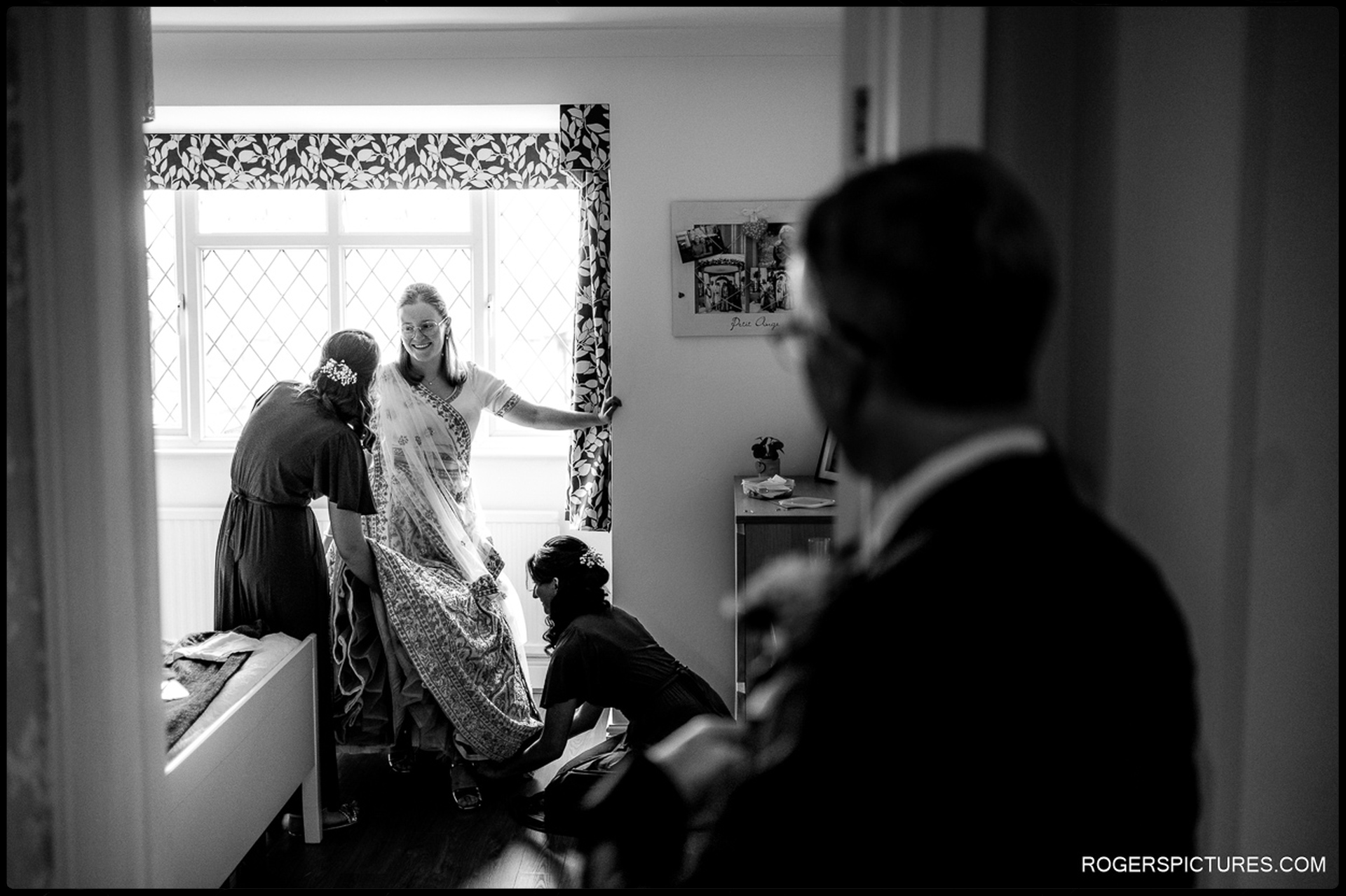 Bride laughs as her bridesmaids adjust her dress while her father watches from the doorway.