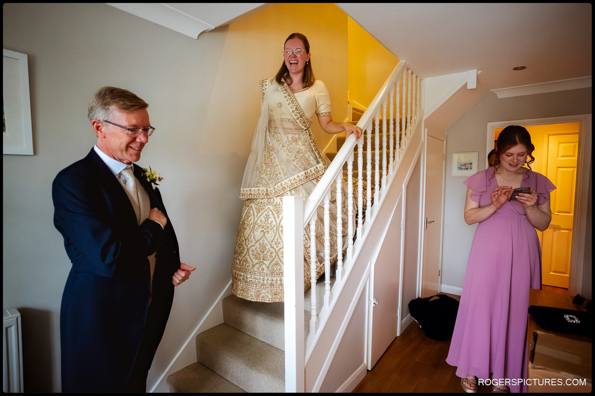 Bride walks downstairs in her gold and cream outfit as her father smiles and a bridesmaid checks her phone.
