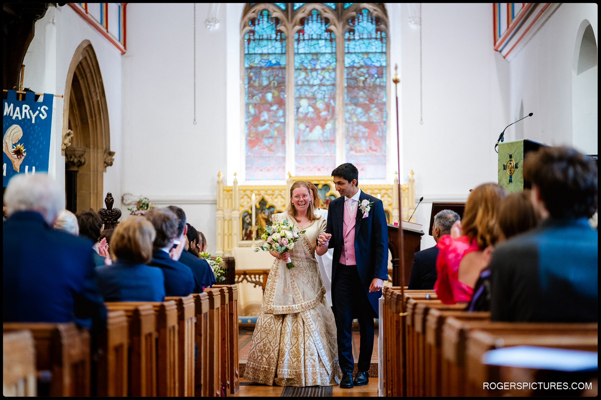 Bride and groom walk back down the aisle smiling as guests applaud inside St Mary’s Church.