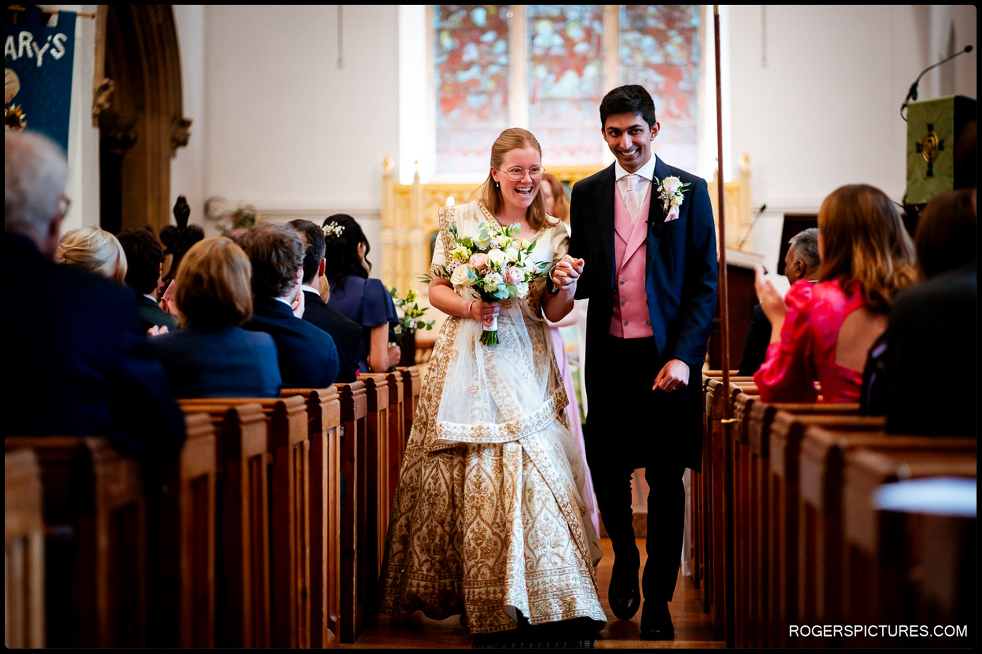 Couple walk arm in arm down the aisle, both smiling as guests look on from the pews.