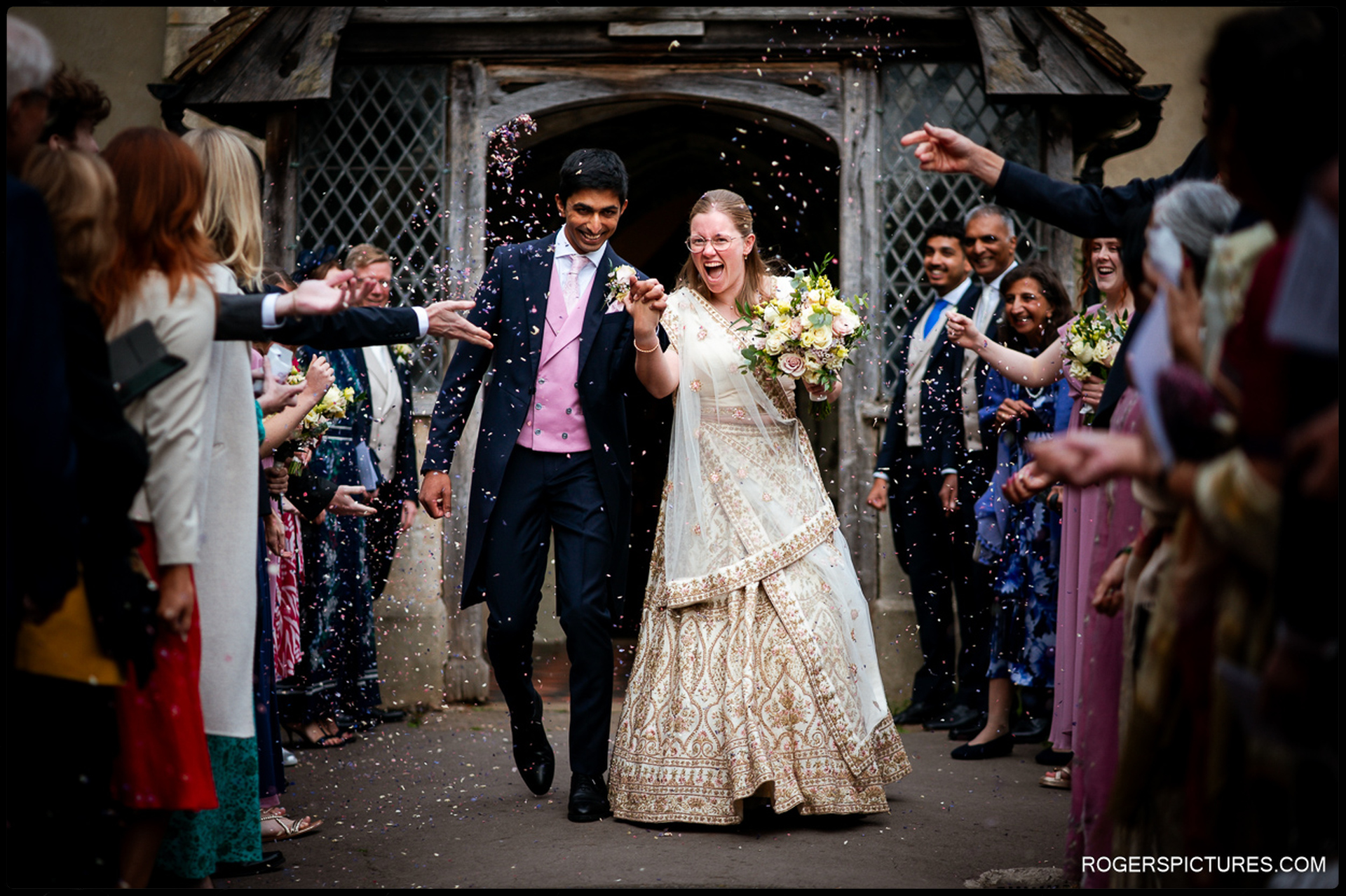 Confetti is thrown over the bride and groom as they walk out of the church laughing.