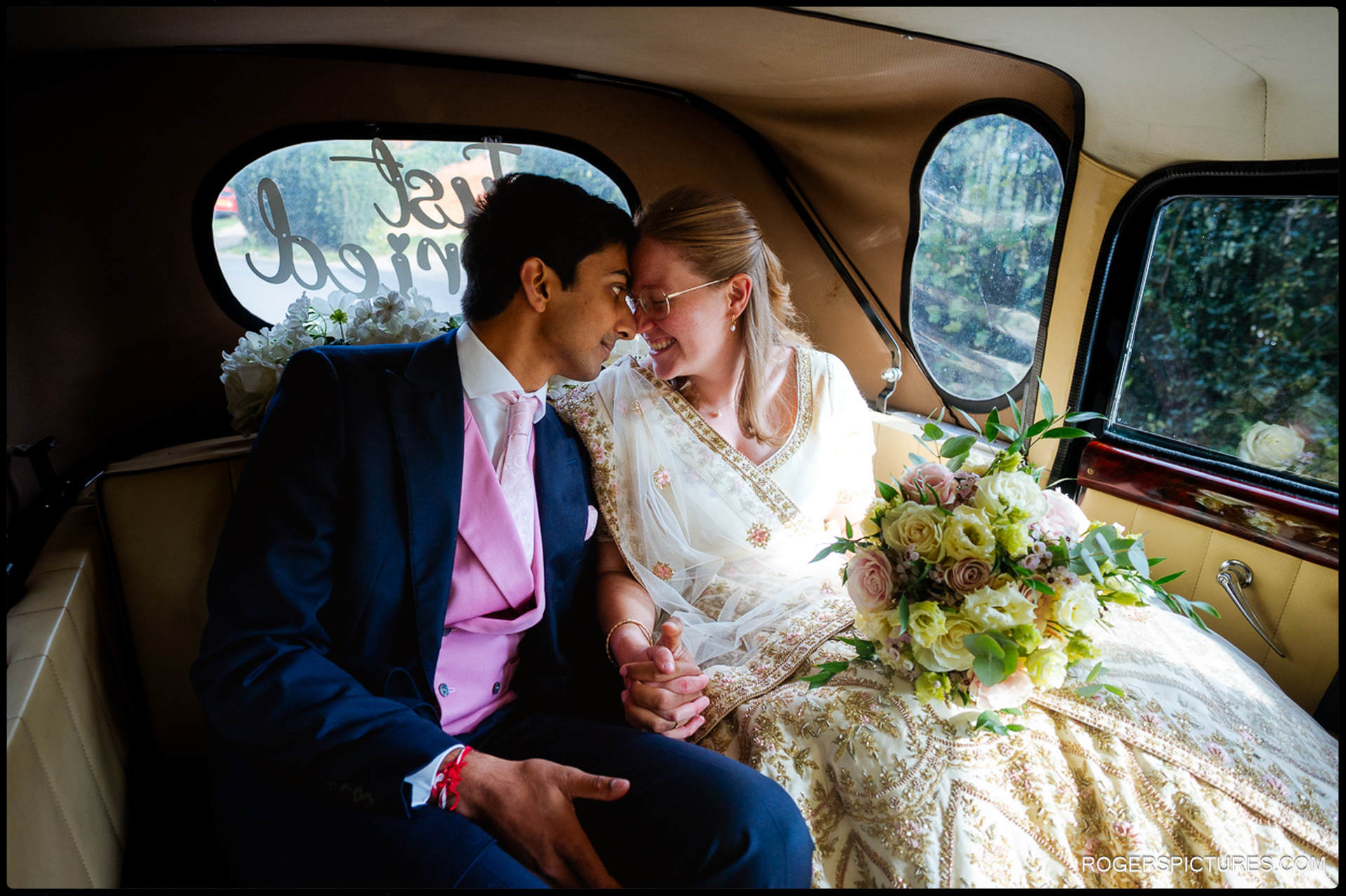 Bride and groom sit close together in the wedding car, holding hands and touching foreheads.