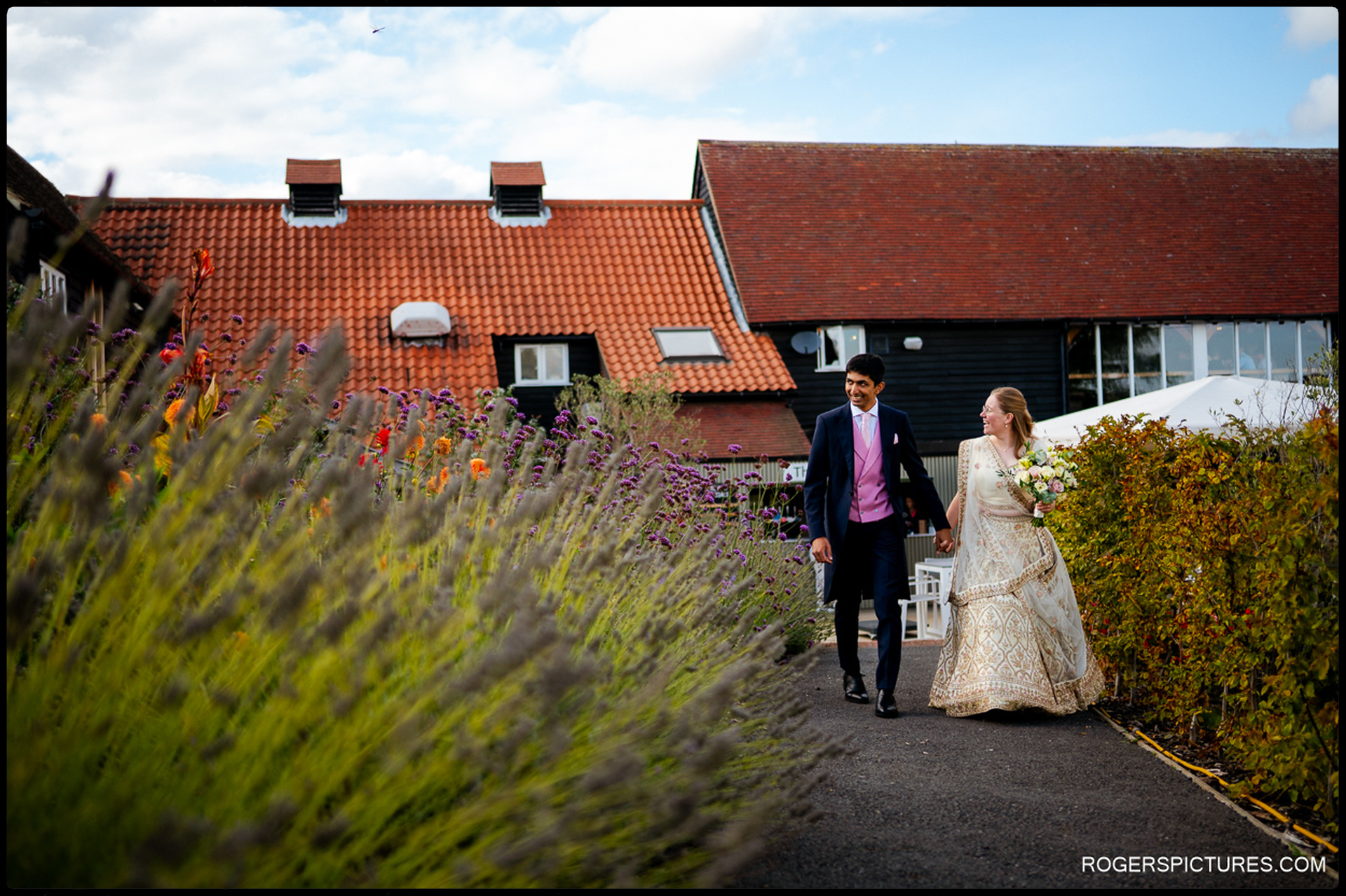 Bride and groom walk along a garden path at the reception venue, surrounded by colourful flowers.