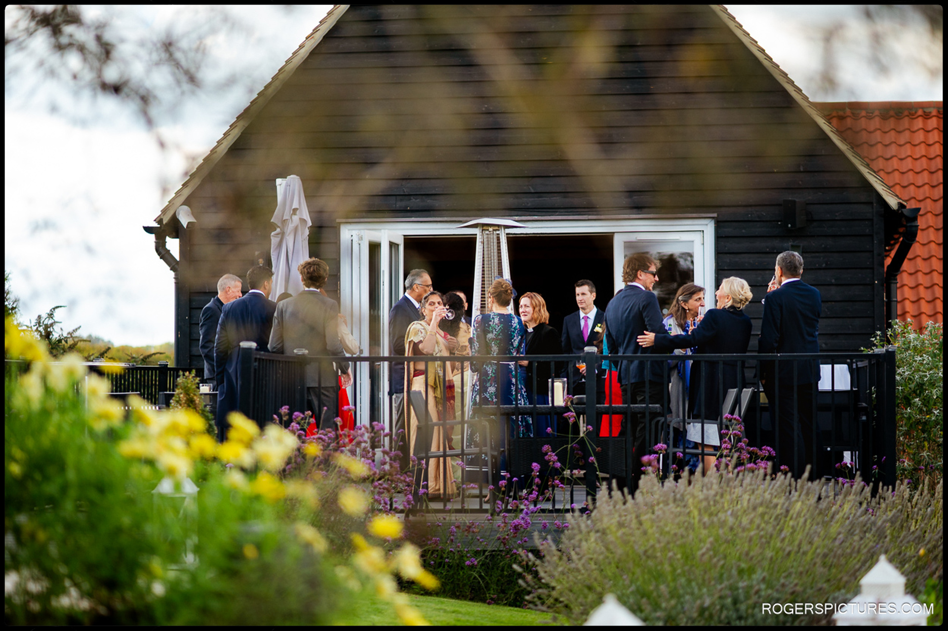 Guests chat and enjoy drinks on the terrace outside the reception barn, seen through garden flowers.