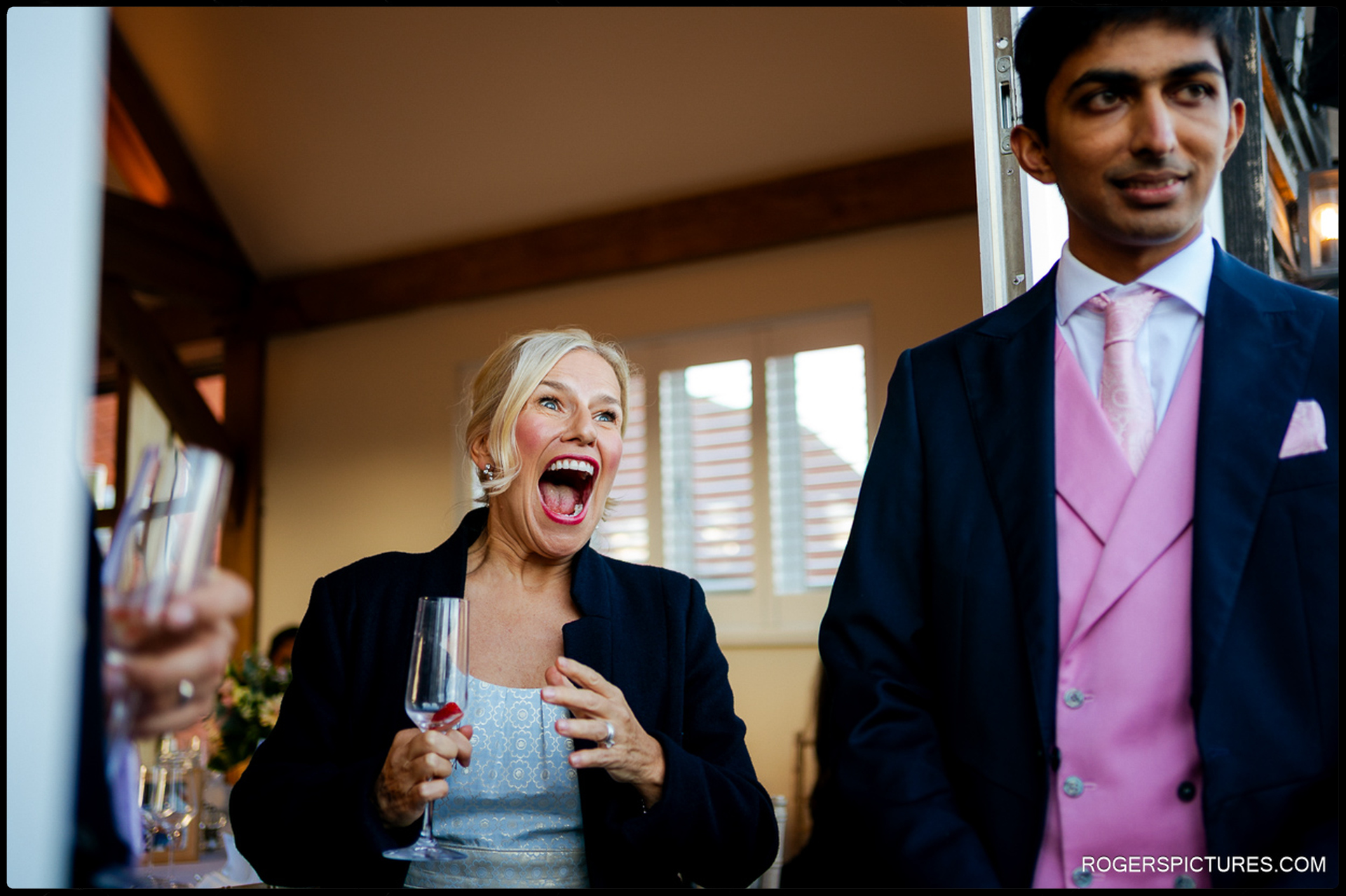 A guest reacts with a wide, joyful expression while holding a champagne flute beside the groom.