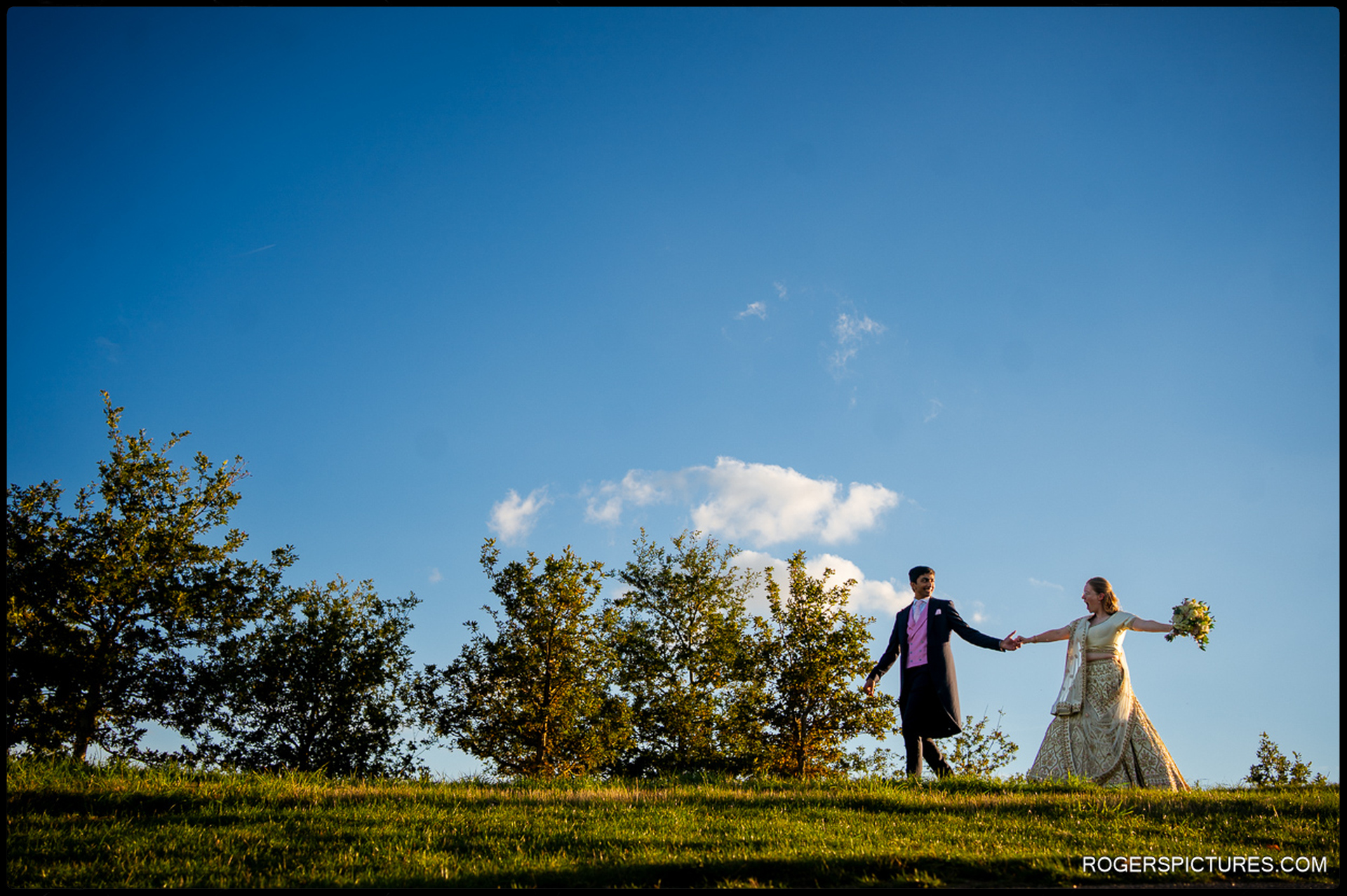 Bride and groom walk hand in hand along the ridge in bright evening light under a clear blue sky.