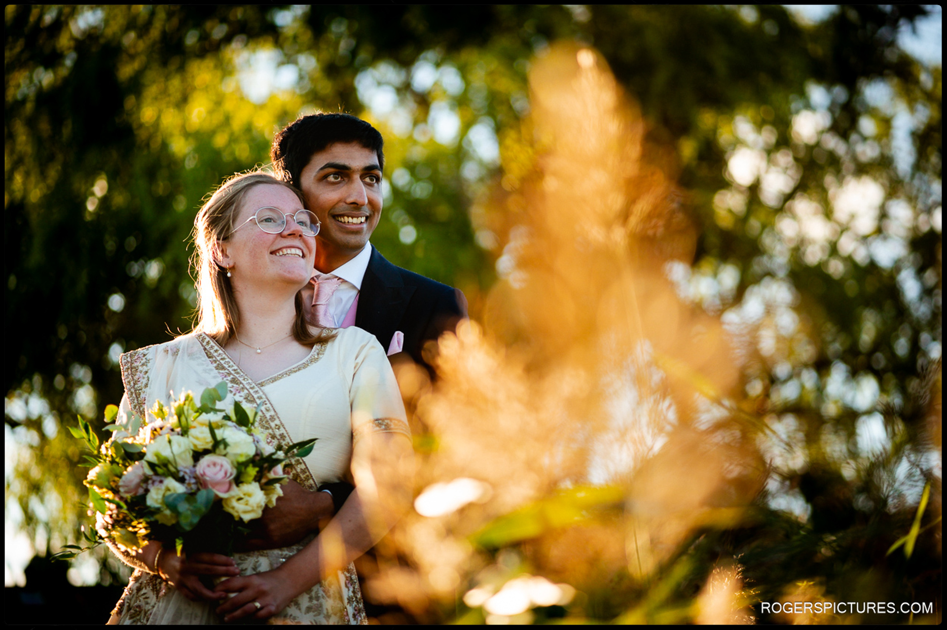 Couple stand together in warm golden light, smiling as they hold each other among garden foliage.