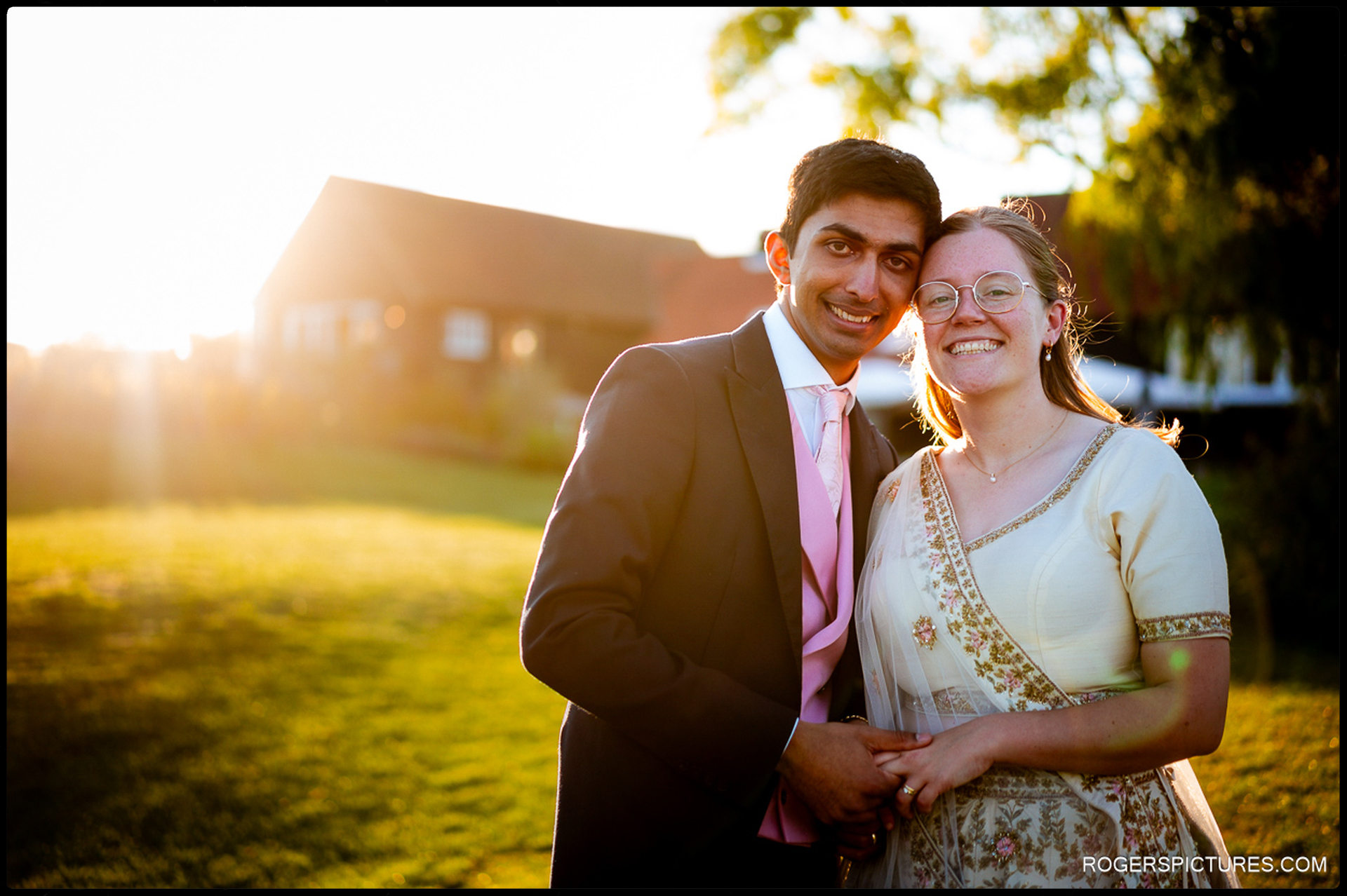 Bride and groom smile at the camera in soft sunset light on the grass outside the venue.