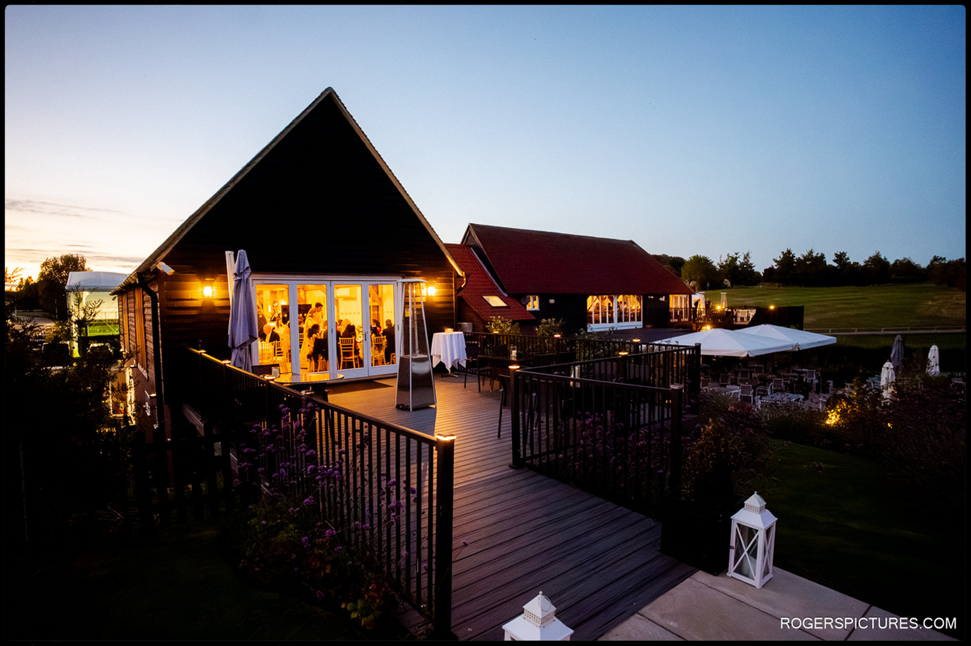 Evening view of the Heron Country Club terrace and barn, lit warmly as guests dine inside.