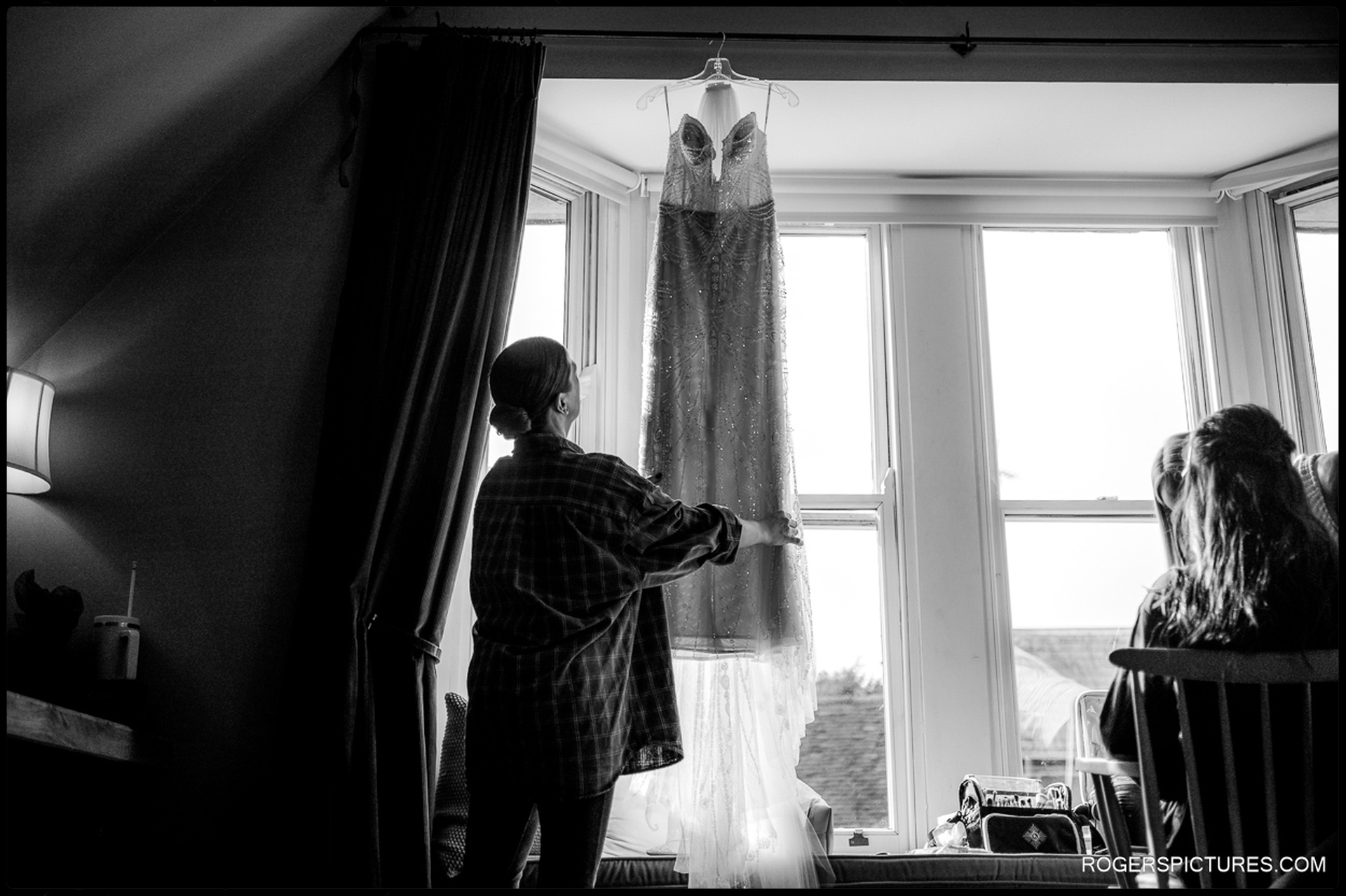 Black and white documentary photo of a bridesmaid adjusting Ciara's wedding dress as it hangs in the sunlit window during bridal prep at The Bell in Ticehurst.