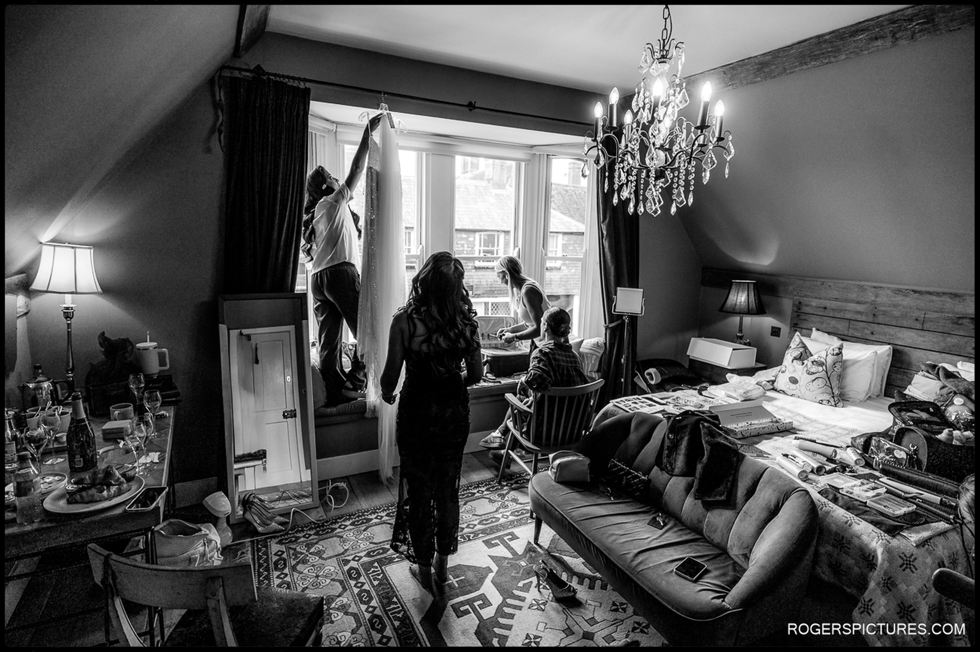 Wide-angle documentary view of the busy bridal preparation room at The Bell in Ticehurst, showing the wedding dress hanging in the window and the unique hotel décor.