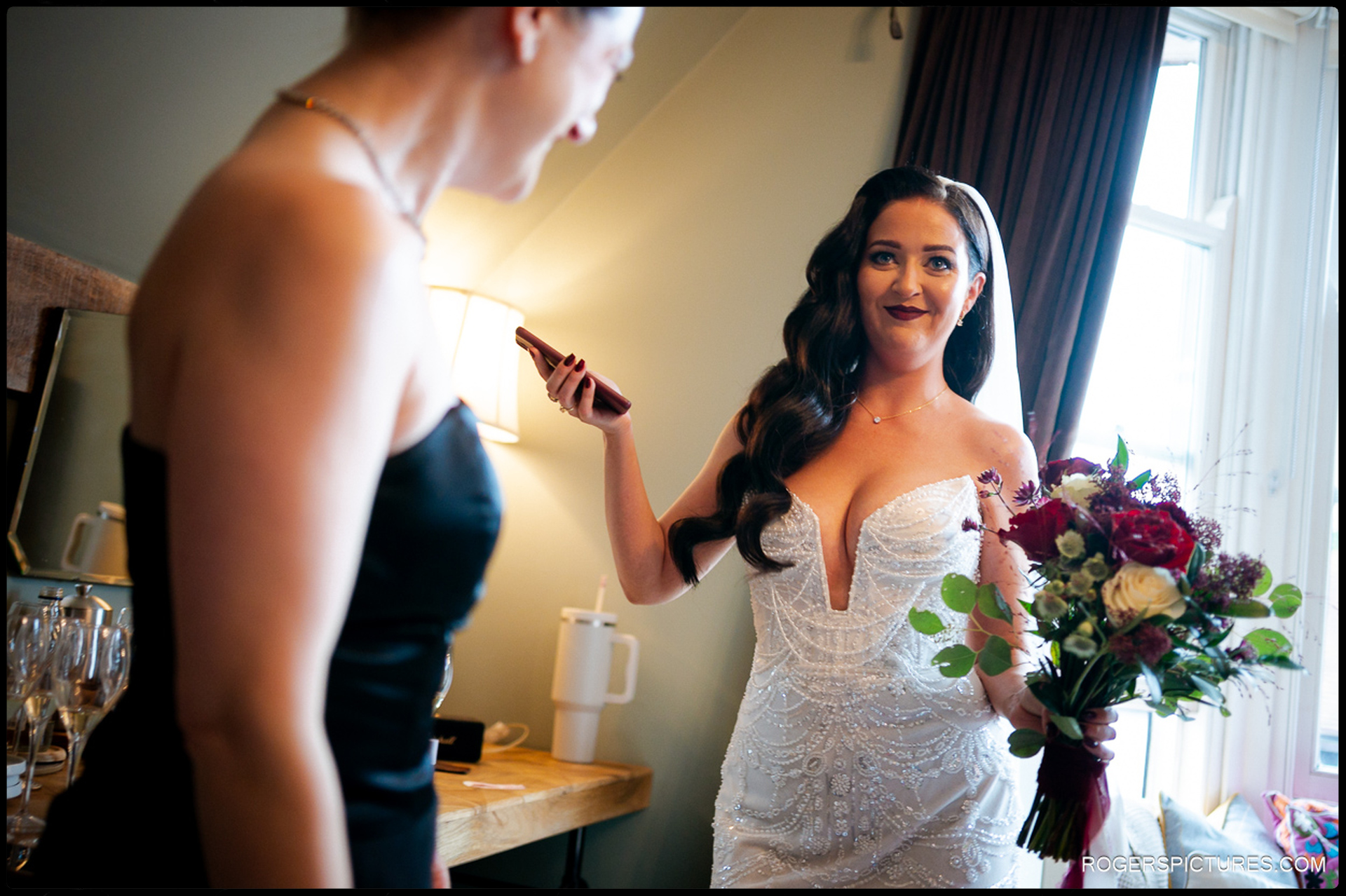 Bride Ciara, holding her deep red and burgundy bridal bouquet and a mobile phone, smiling while talking to a bridesmaid during prep at The Bell in Ticehurst.