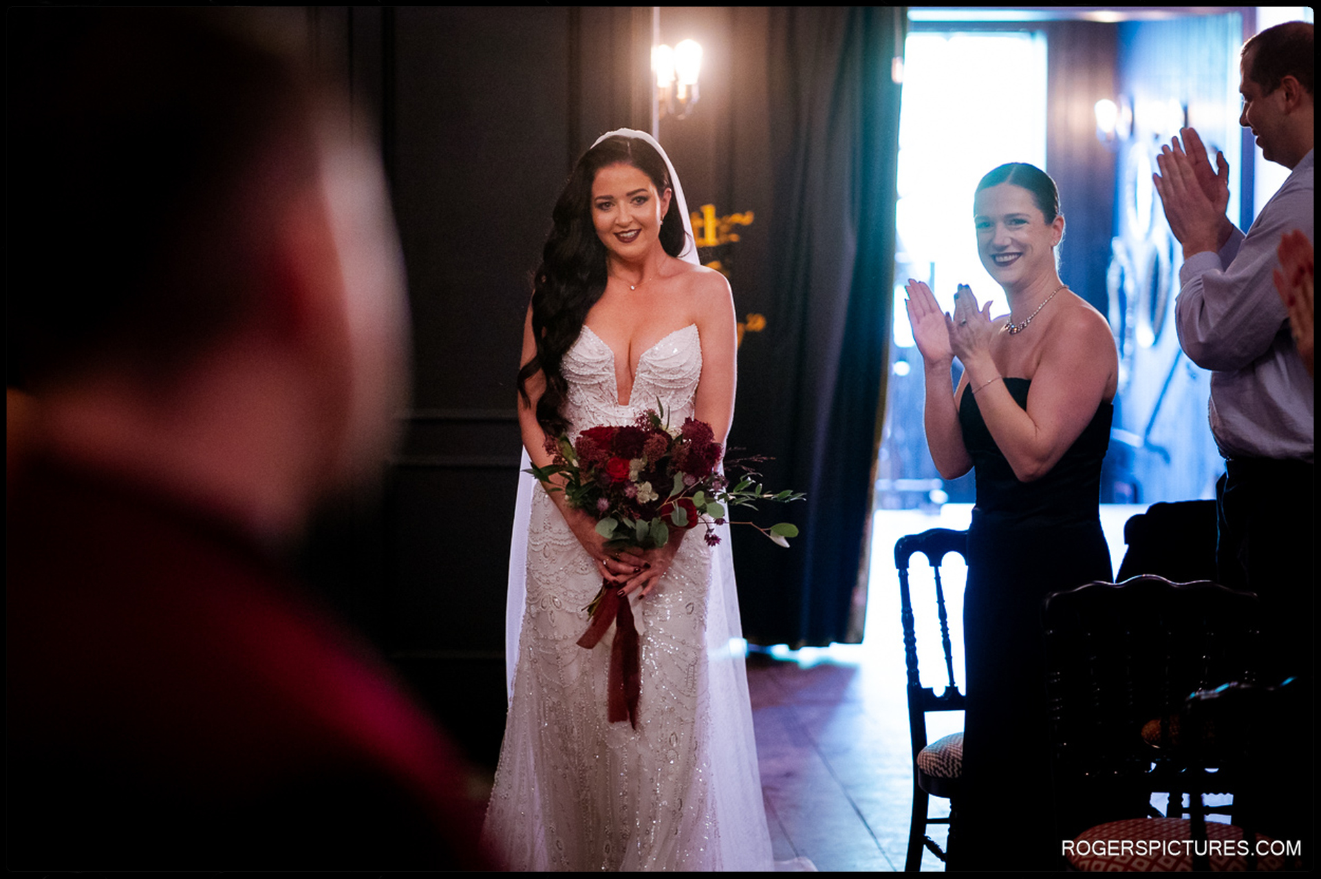 Bride Ciara smiling as she enters the ceremony space, holding her bouquet while guests applaud, captured with documentary lighting at The Bell in Ticehurst.
