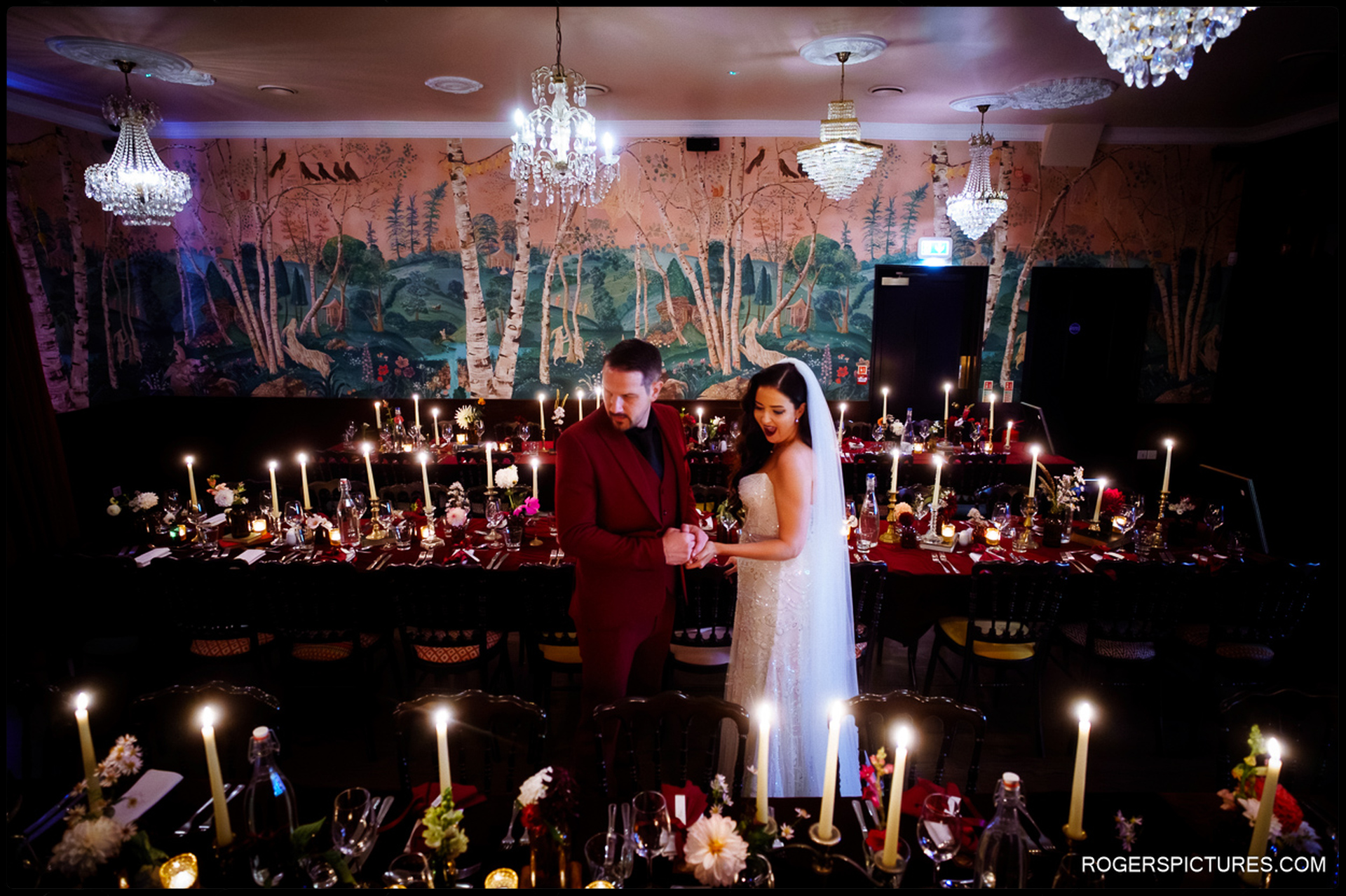 The couple posing in the candlelit dining room, featuring the distinctive hand-painted wallpaper and chandeliers.