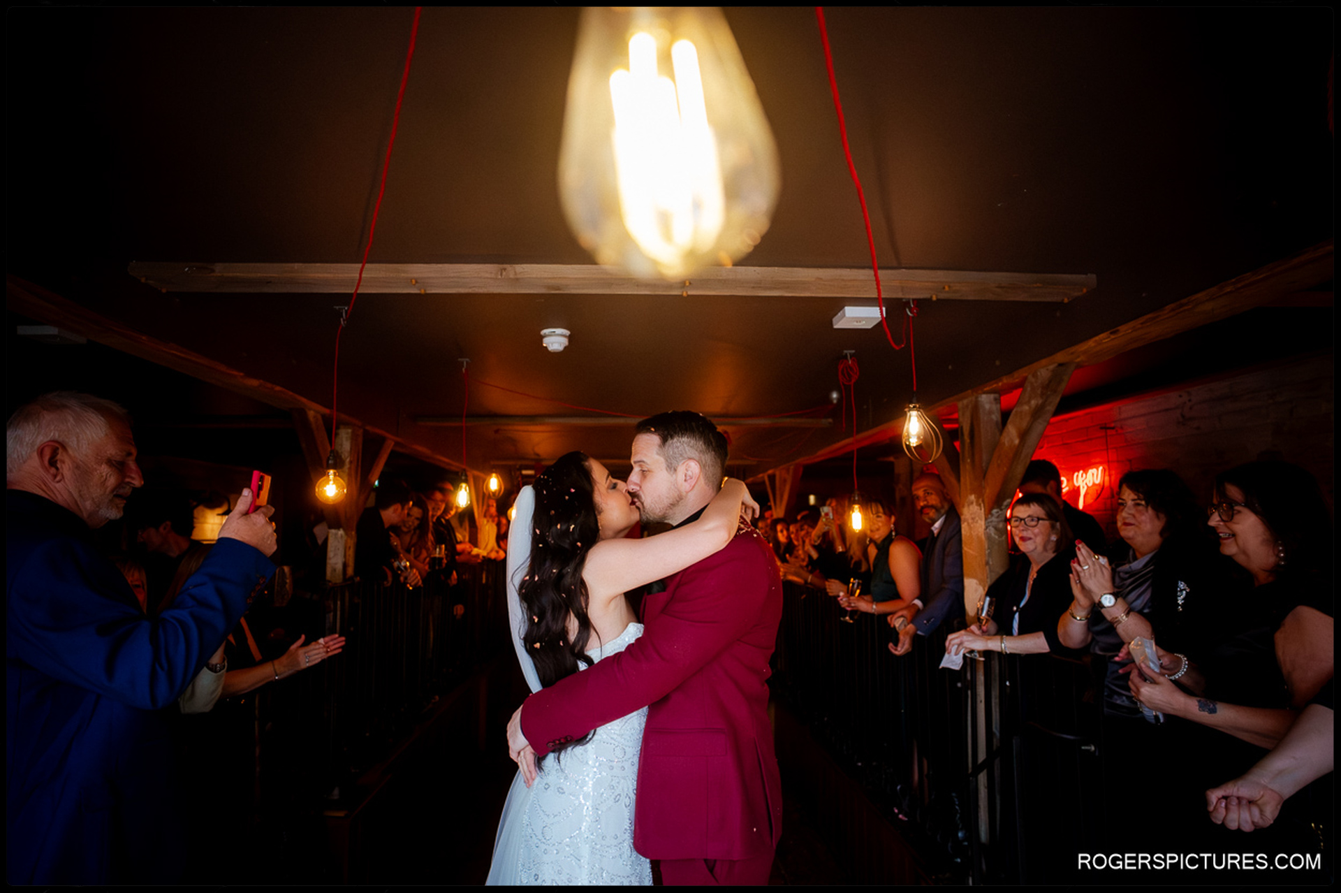 A romantic close-up of the couple kissing under a single Edison bulb, surrounded by cheering guests during the evening reception.