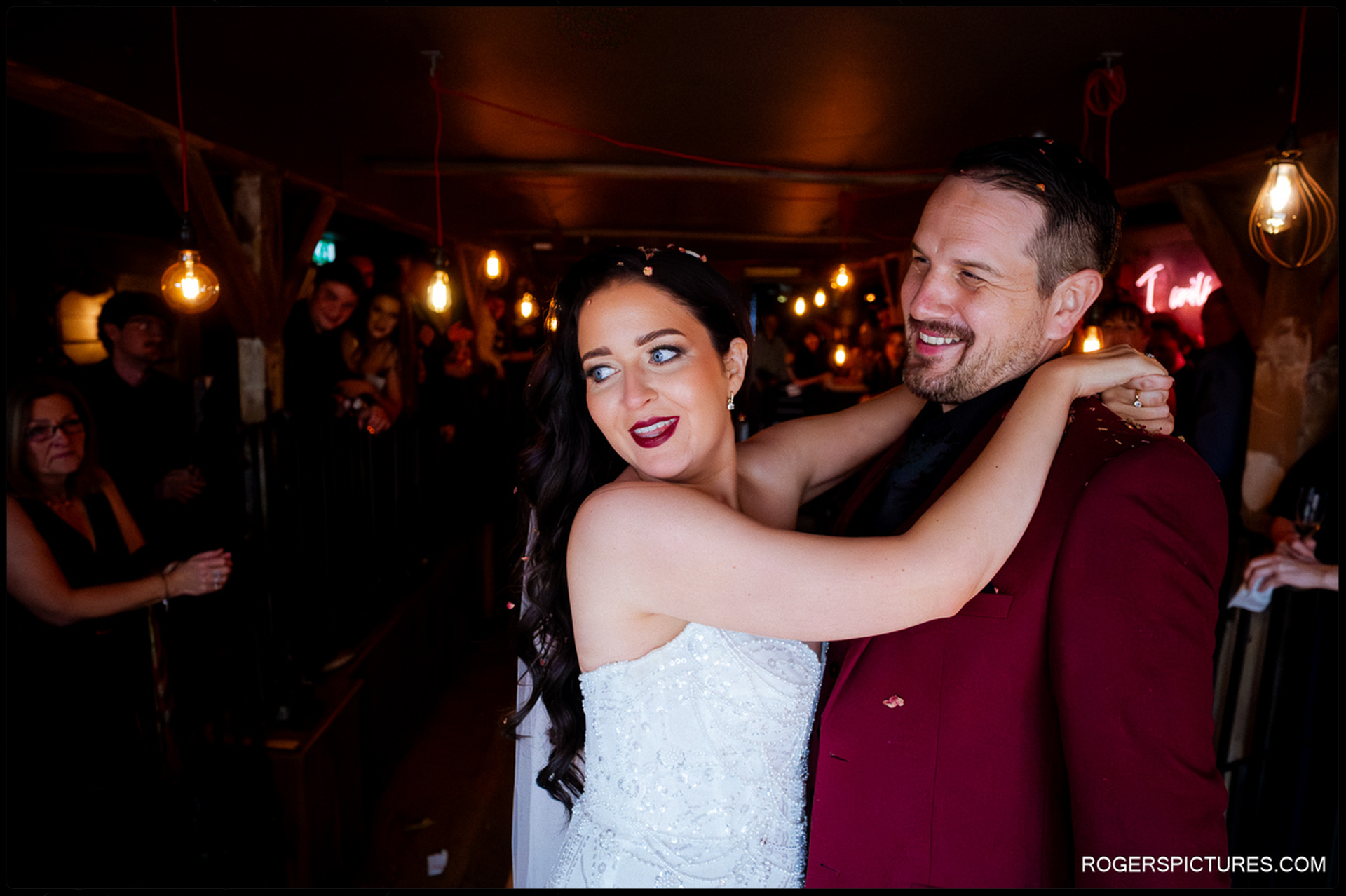 The couple embracing and smiling during the First Dance, surrounded by warm, dark lighting and exposed bulbs.