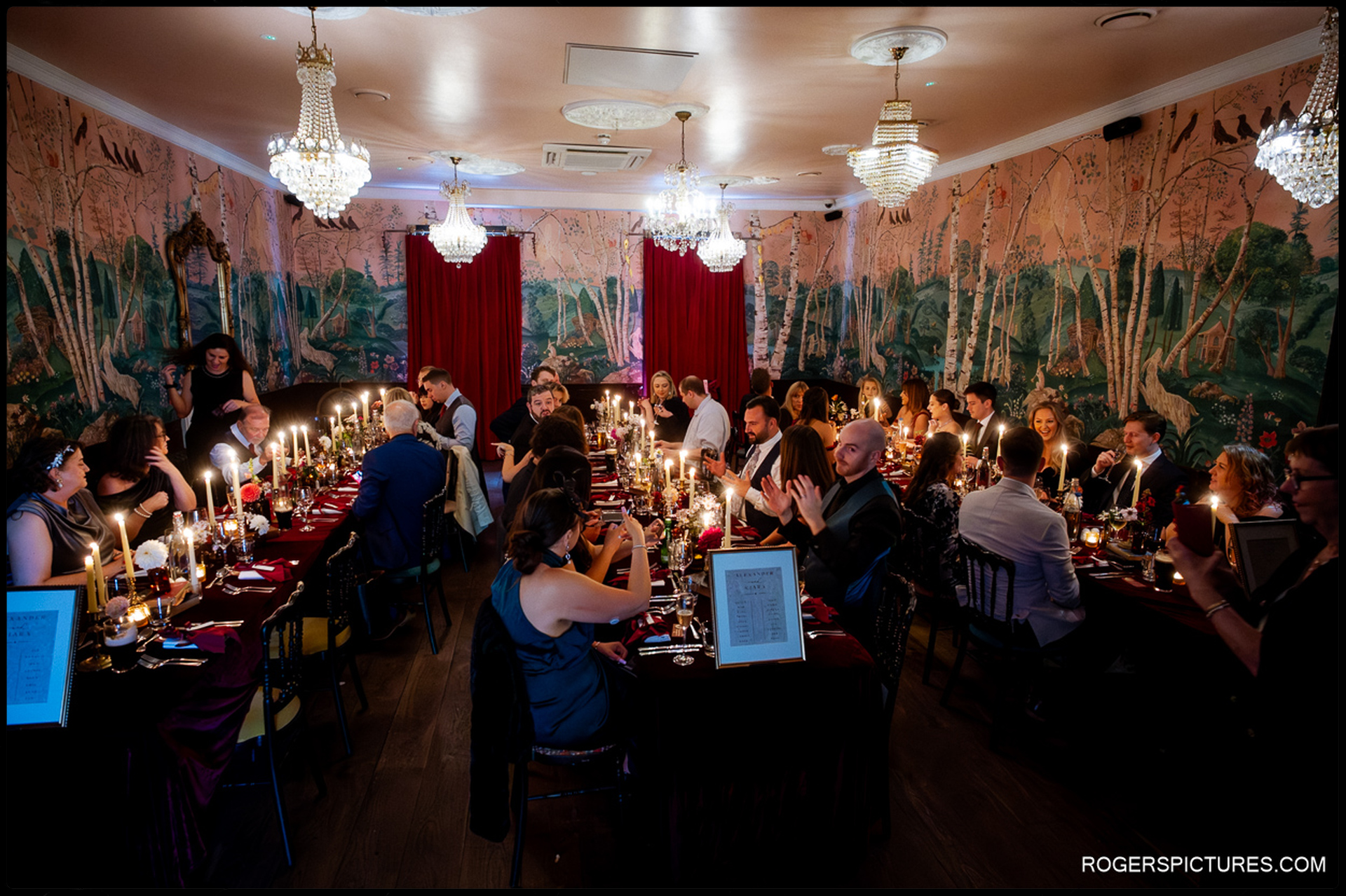 Guests seated at the long, candlelit wedding breakfast tables, surrounded by the distinctive forest mural wallpaper.