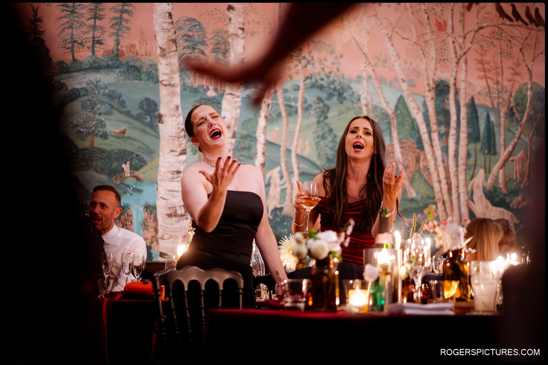 Two guests cheering loudly and raising their hands at the wedding breakfast table during the evening speeches or toasts.
