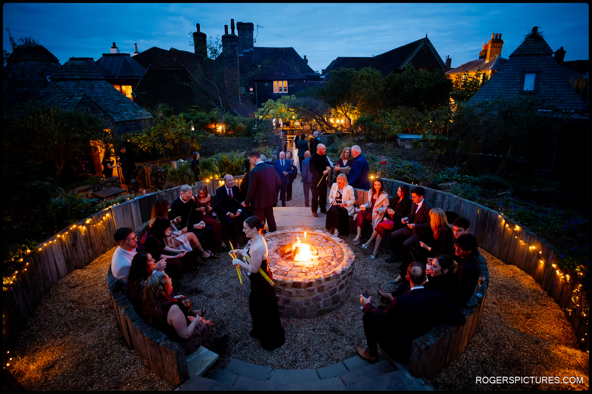 Wide shot of the wedding guests gathered around the outdoor fire pit at dusk, creating a warm, illuminated social area in the garden.