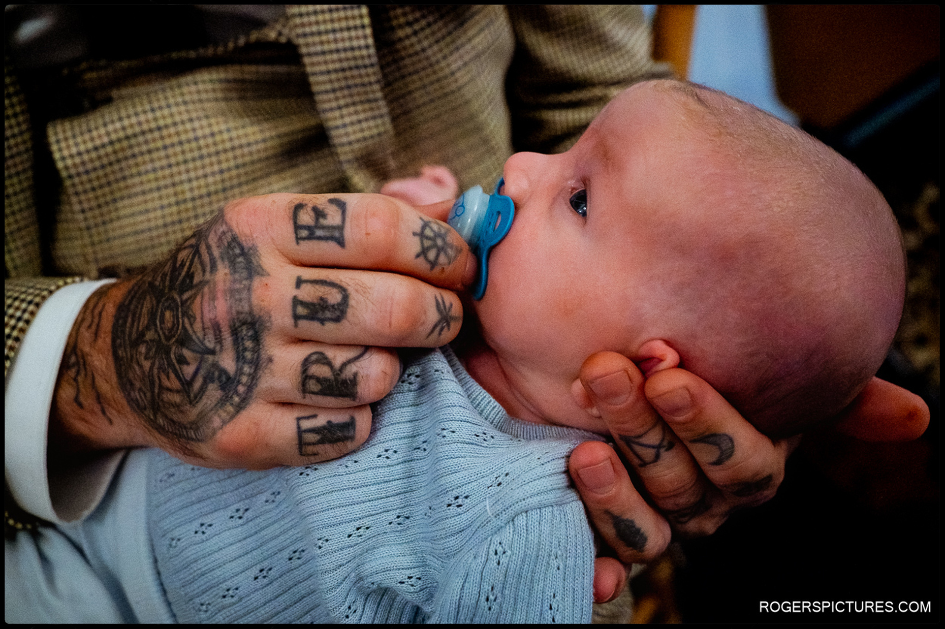Tattooed hands gently holding a baby with a pacifier during the wedding at Rumbolds Farm.