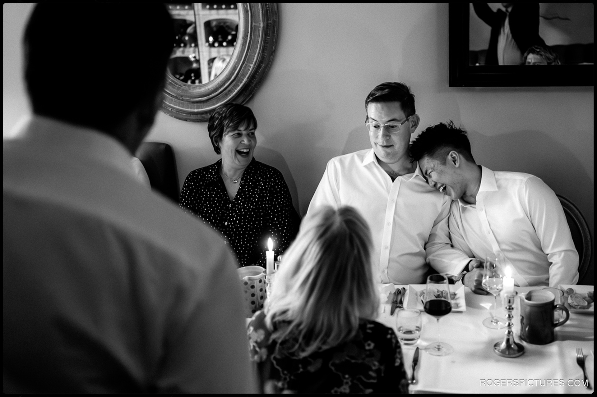 A black and white documentary photo of a couple and their guests laughing together at a candlelit dinner table during the wedding breakfast.
