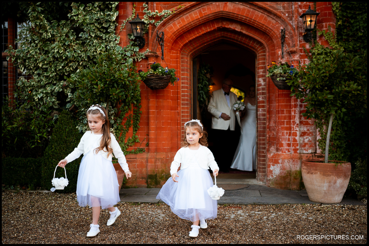 A documentary shot of two flower girls in white dresses walking along a gravel path outside the red brick entrance of Woodhall Manor.