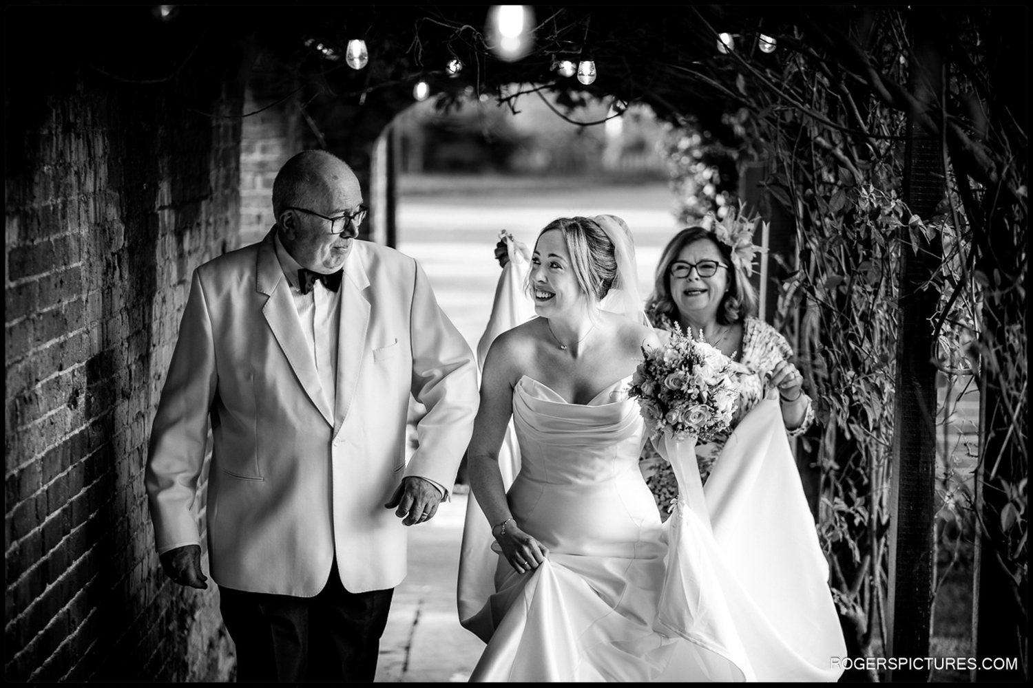 A black and white candid photo of a bride laughing while walking through a festoon-lit archway with her father and another guest.