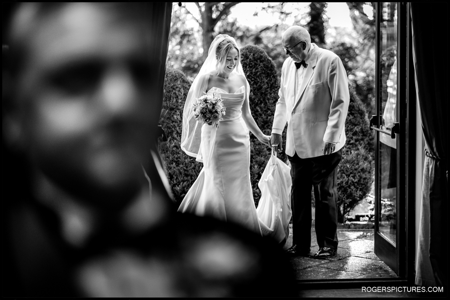 A black and white documentary photograph of a bride and her father holding hands as they prepare to enter the ceremony room at Woodhall Manor.
