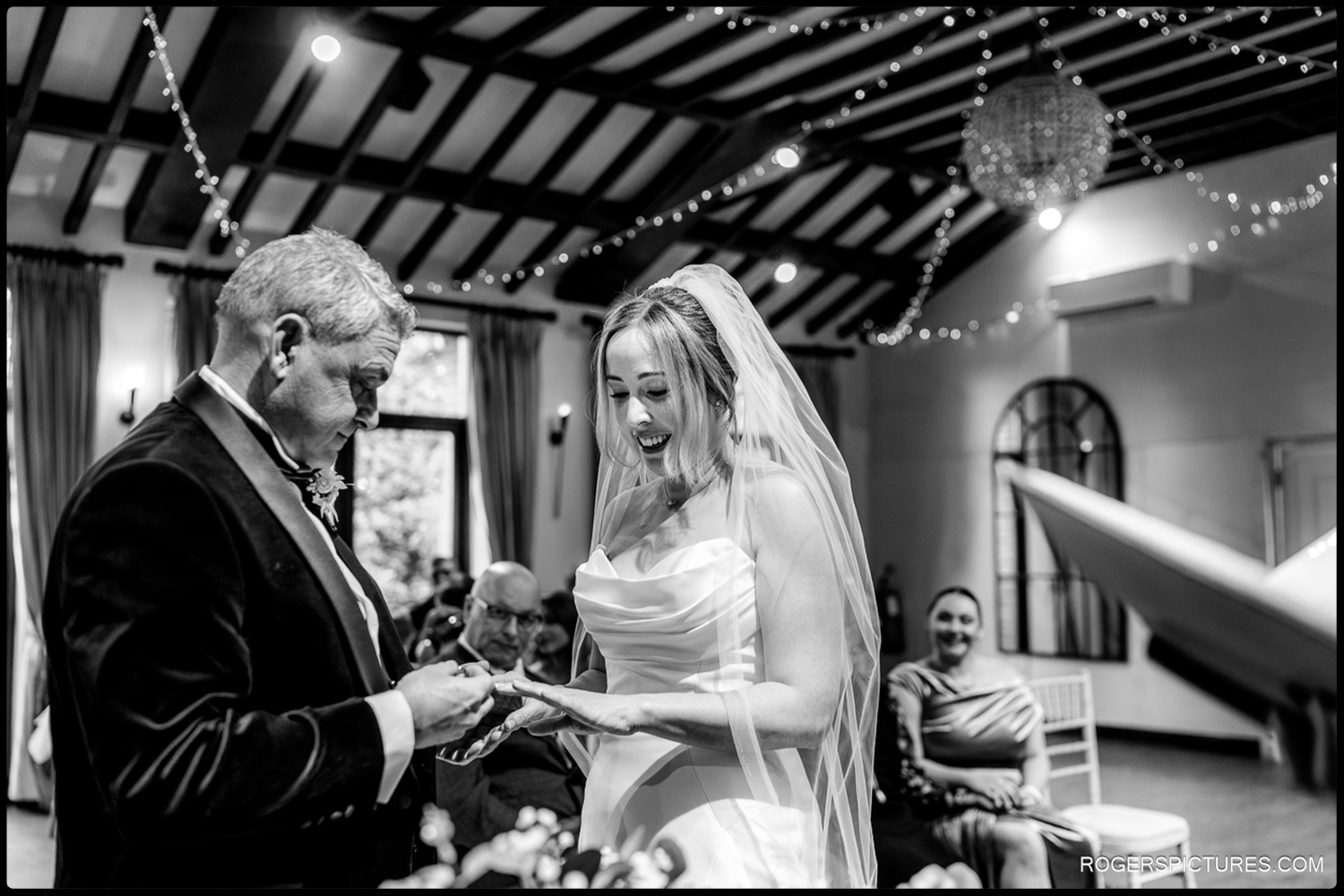 A black and white candid photo of a bride laughing as the groom places a ring on her finger during their indoor wedding ceremony.