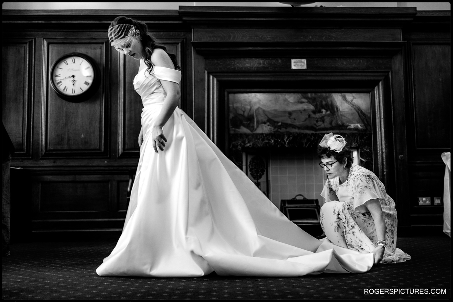 A documentary photograph of a bride standing in her white wedding gown in front of a historic fireplace, while an assistant carefully adjusts her train.